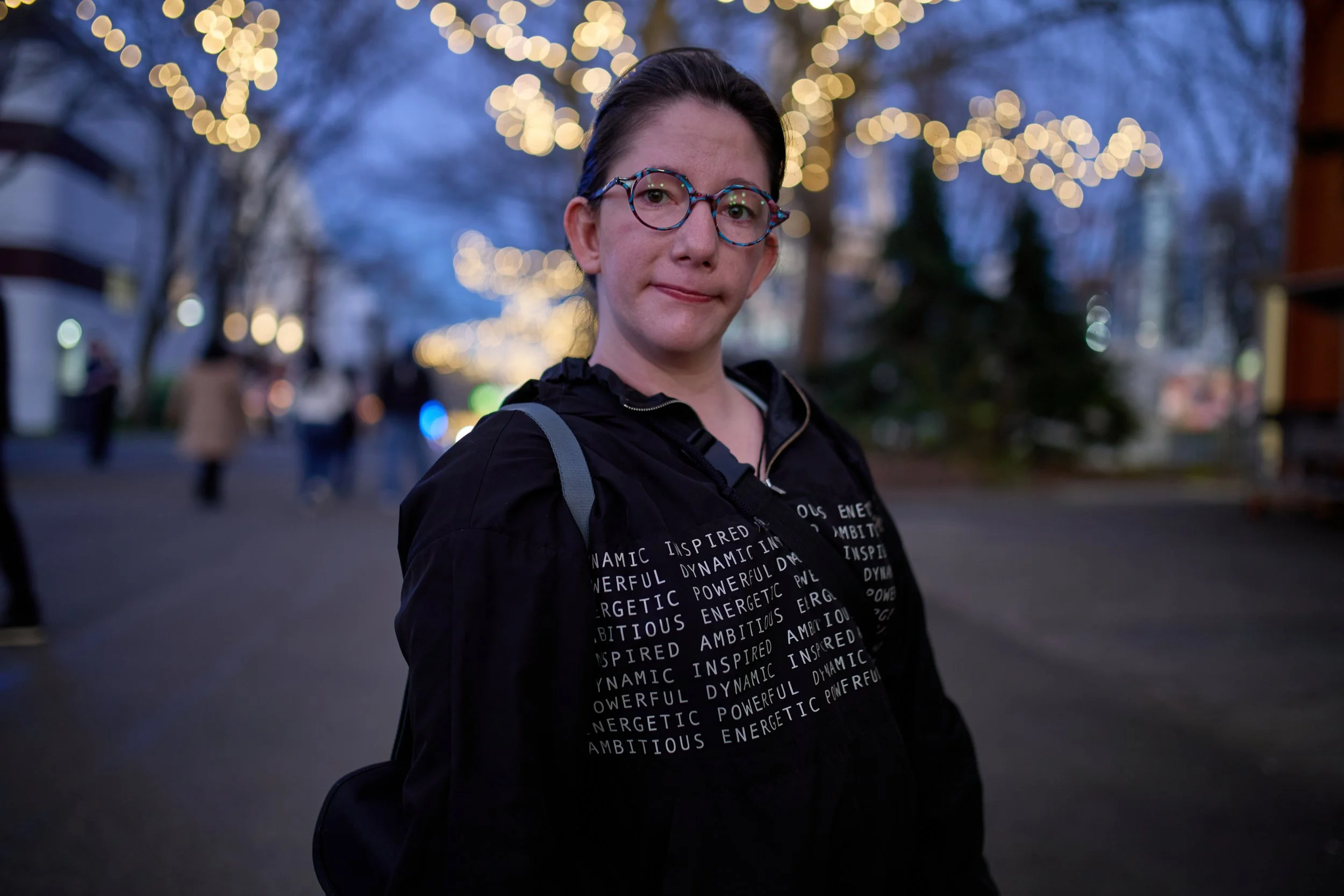 A young woman with dark hair and glasses standing outdoors at dusk with string lights above her, wearing a black jacket with words like 'dynamic,' 'inspired,' 'powerful,' and 'energetic' printed on it, with blurred people and buildings in the background.