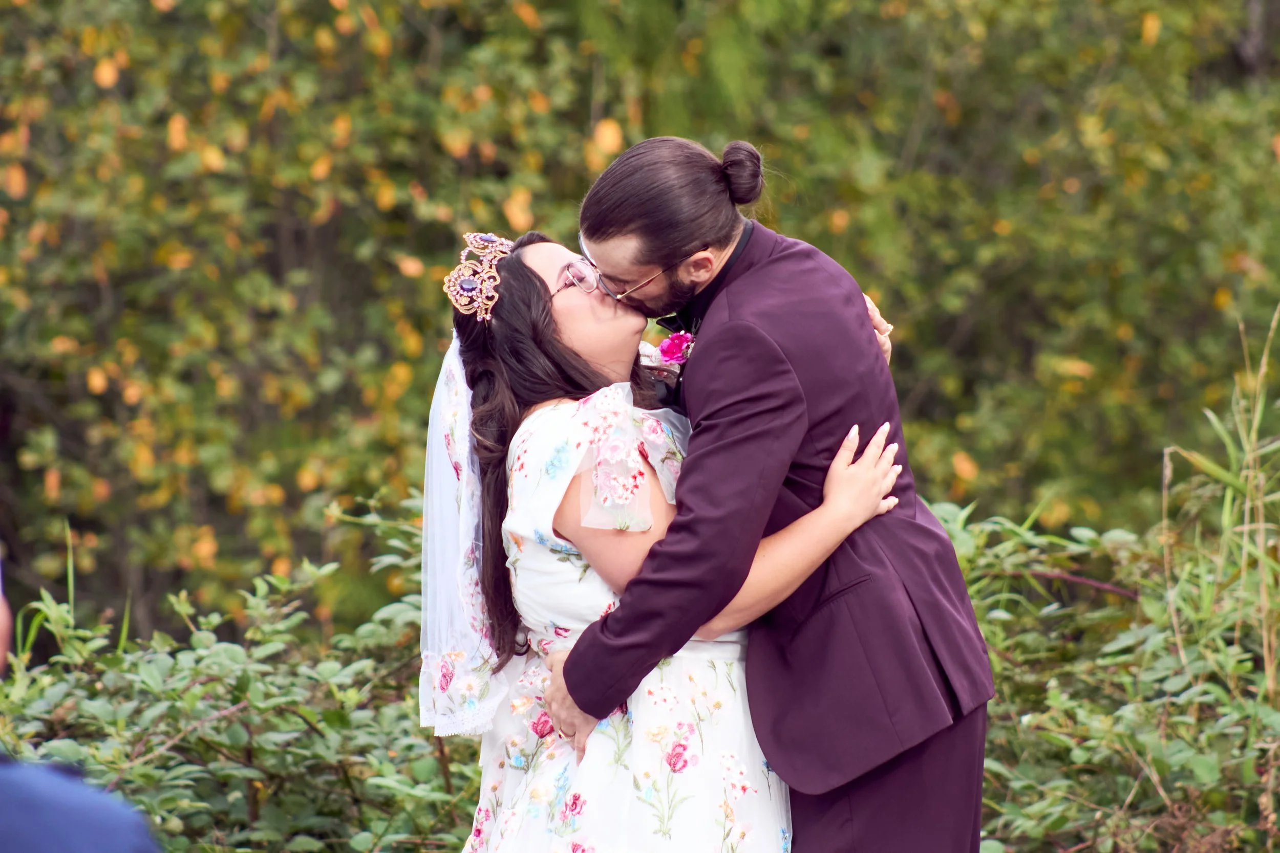 A couple in wedding attire sharing a kiss outdoors in a natural setting with green trees and plants.
