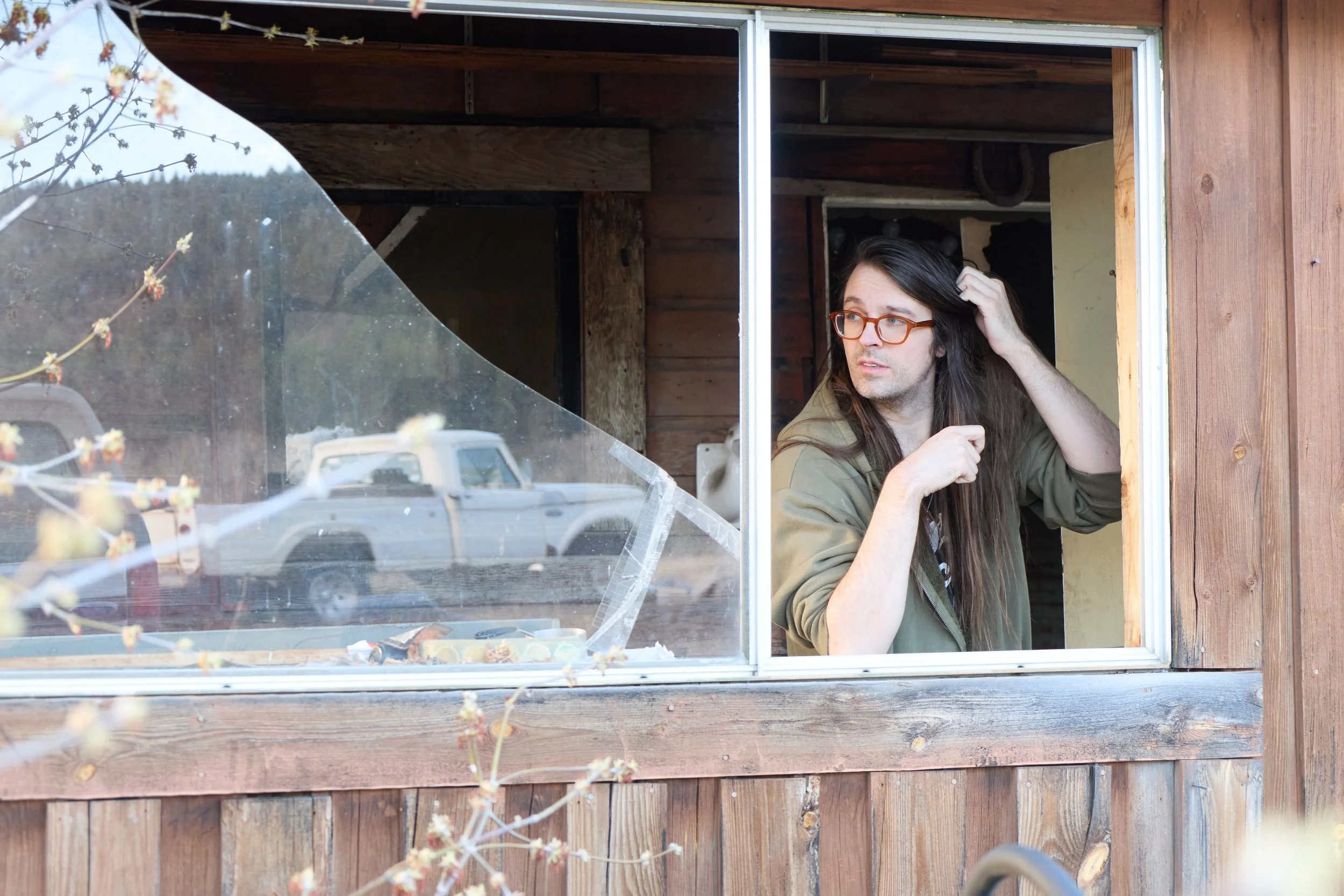 A man with long dark hair, wearing glasses and a green jacket, looks out a window with a surprised or curious expression, scratching his head.