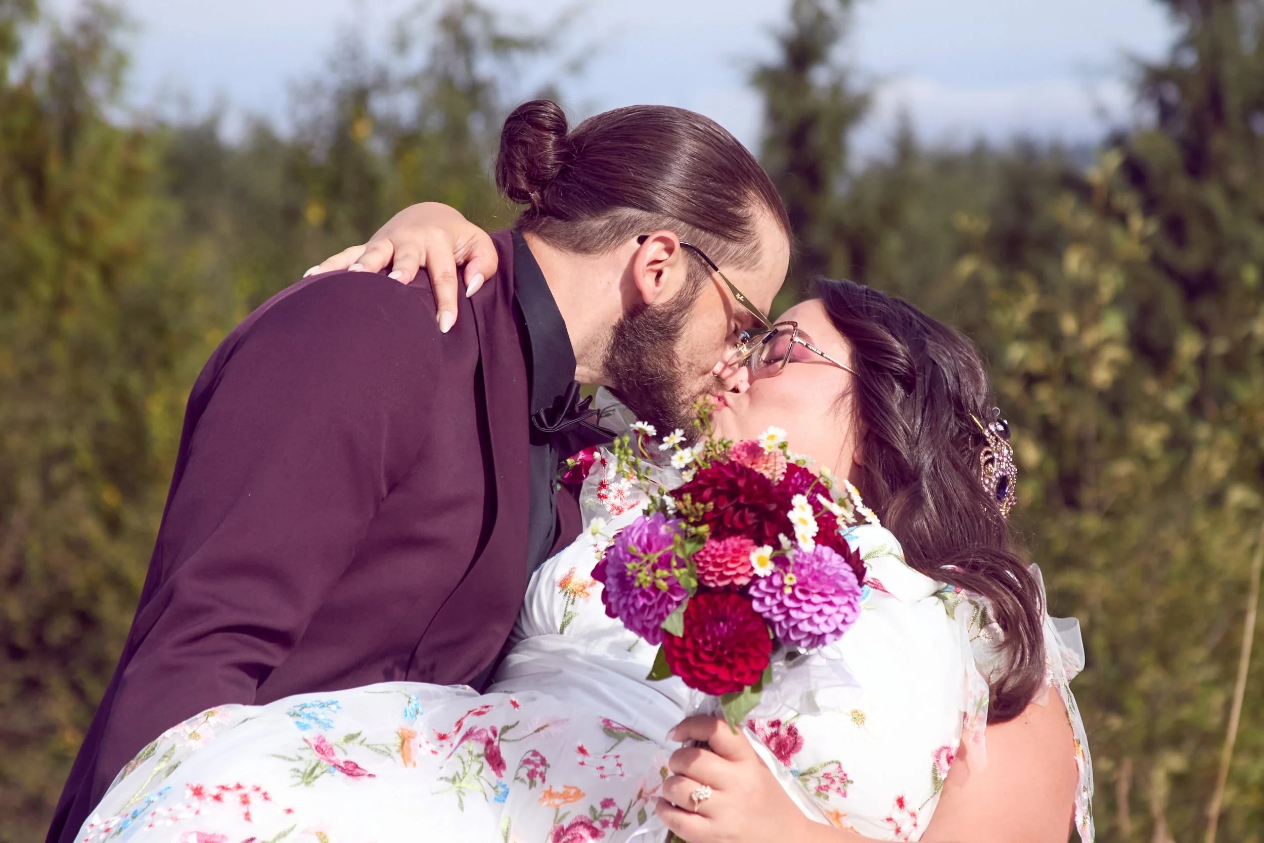 A couple kissing outdoors during the daytime, with the woman holding a bouquet of colorful flowers and wearing a white dress with floral patterns.