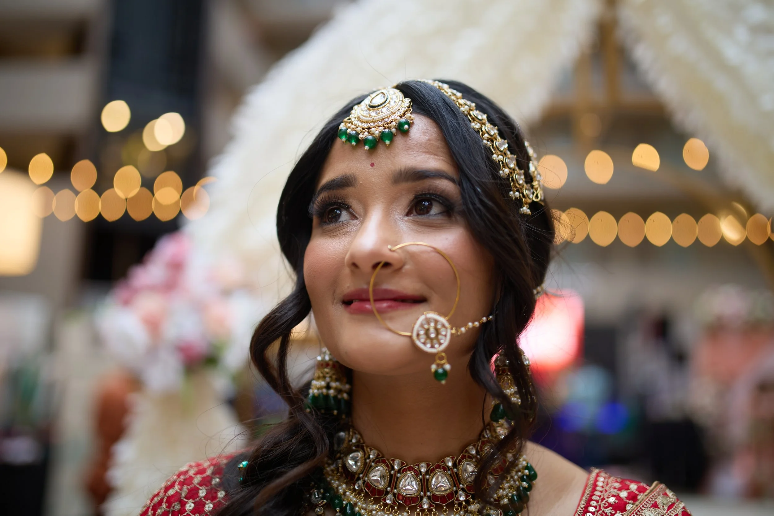 A woman dressed in traditional Indian jewelry and attire, with intricate jewelry featuring green and gold, and a red patterned blouse. The background shows a blurry display of lights and decorative items.