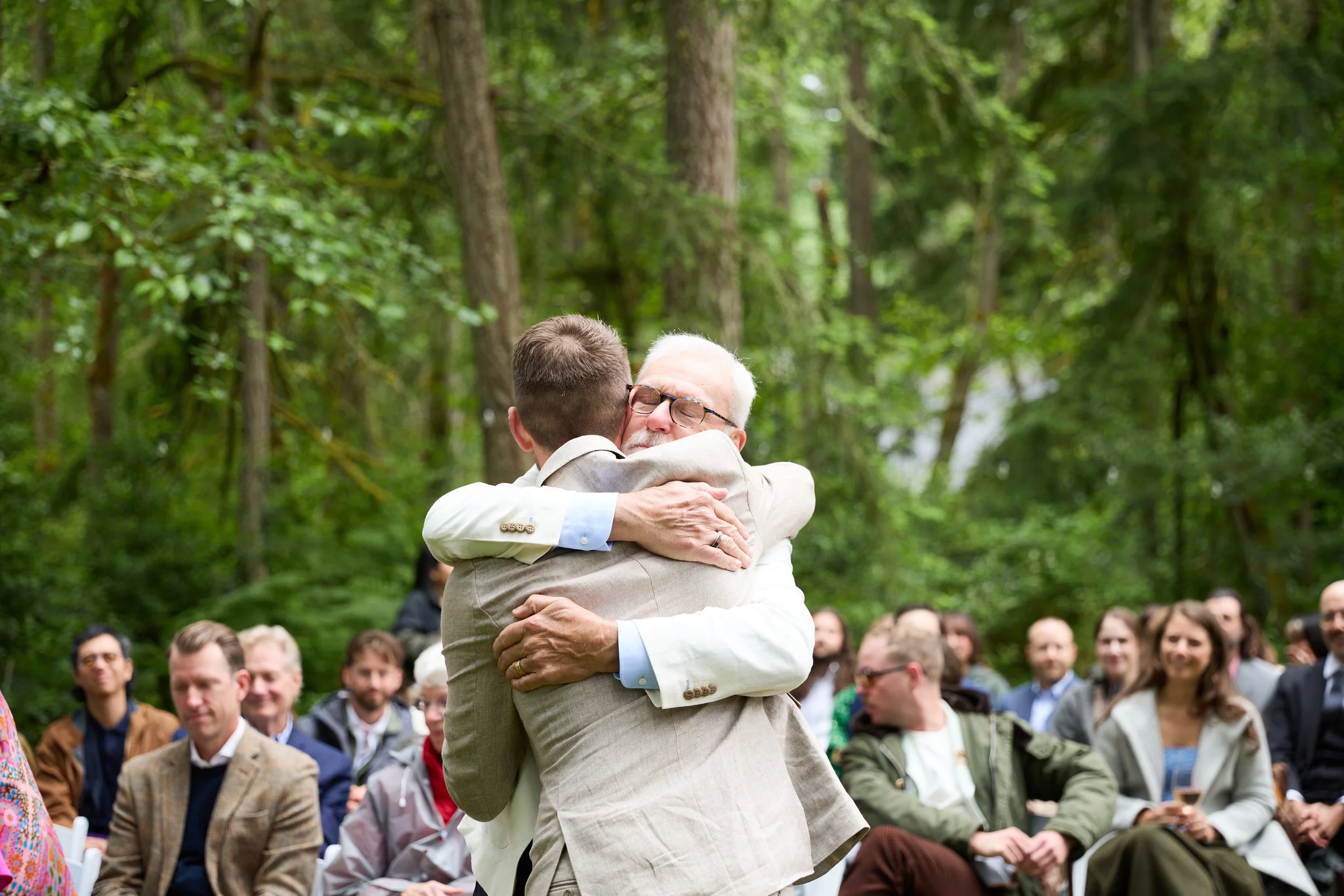 During a wedding, a father and son embracing each other in a hug while a crowd of people watch them outdoors in a wooded area.