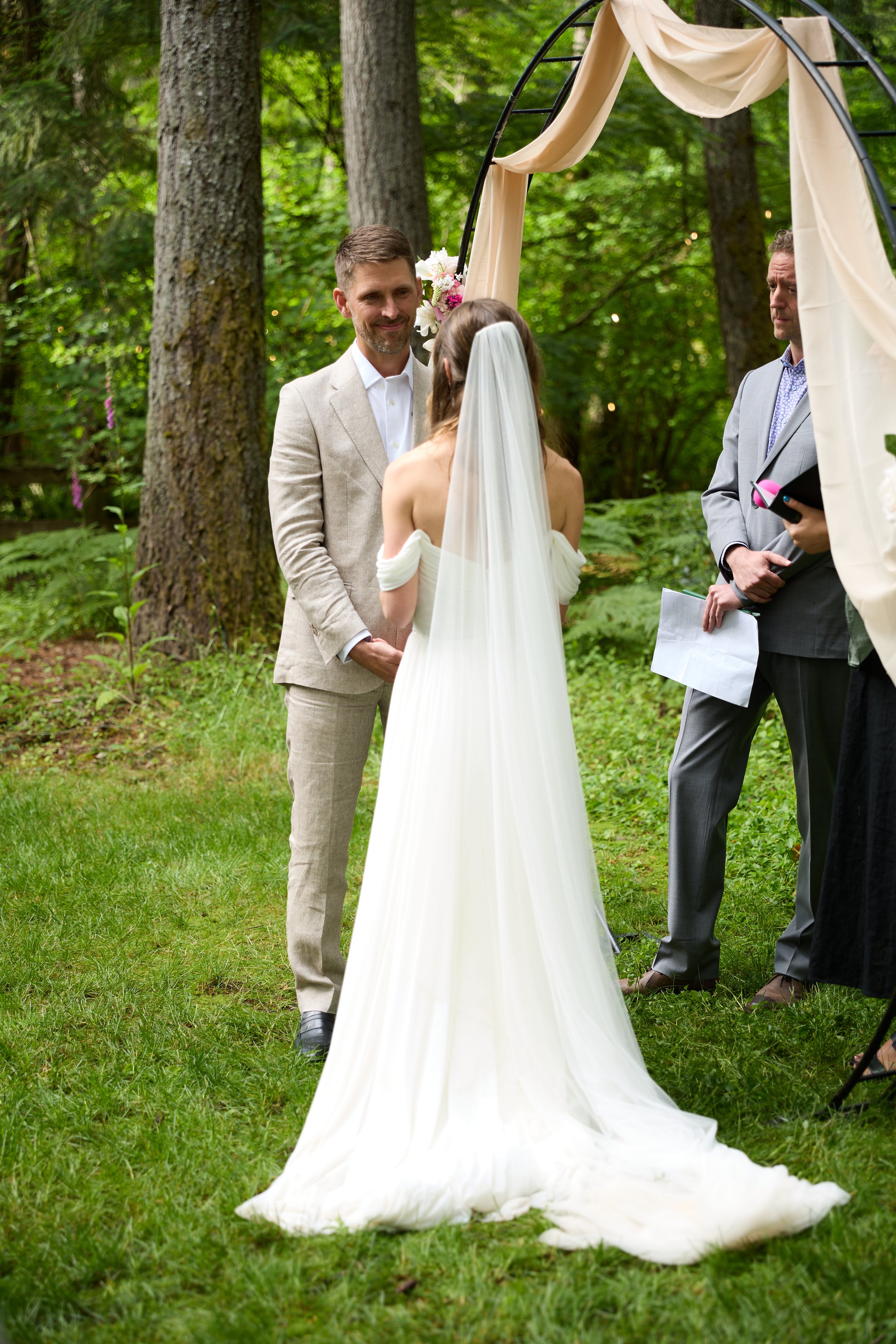 A couple getting married outdoors in a forest, with an officiant standing nearby. The bride is in a white wedding dress with a veil, and the groom is in a light-colored suit. They are standing under a black metal arch with draped fabric and flowers.