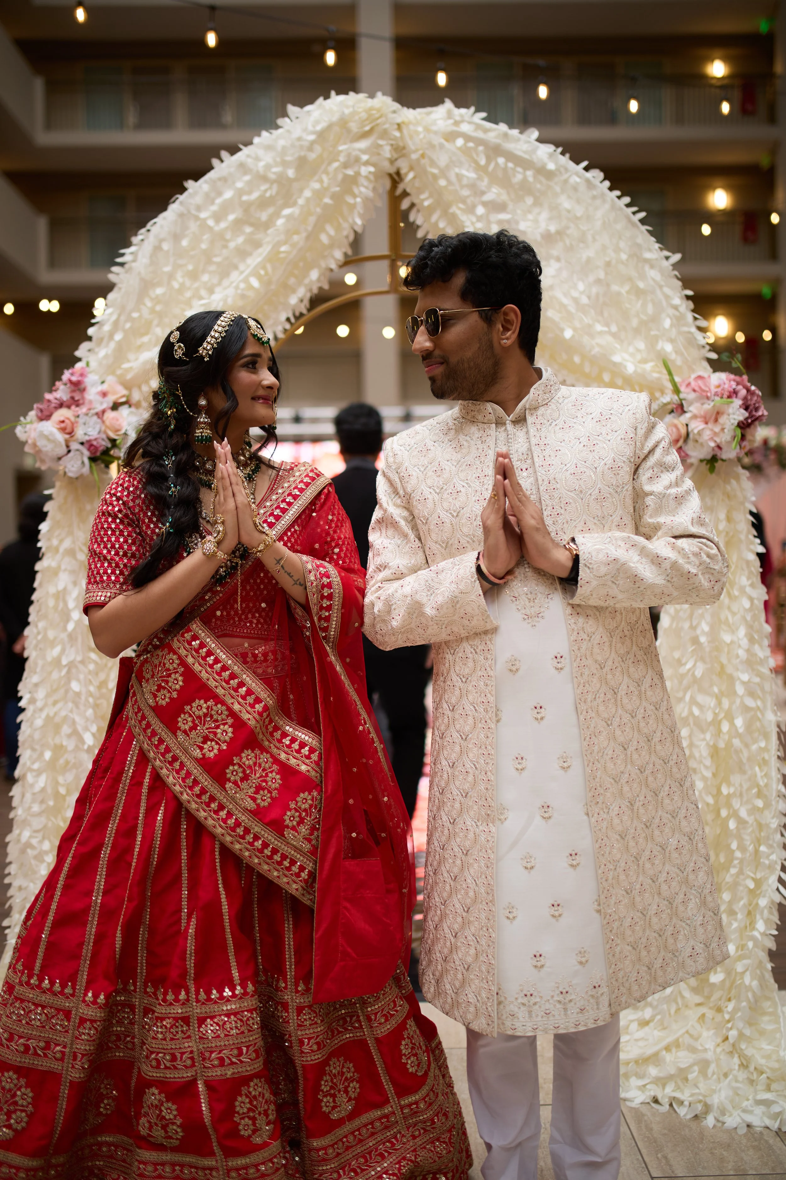 A bride and groom in traditional Indian wedding attire standing under a floral arch, hands pressed together in greeting, smiling at each other indoors.