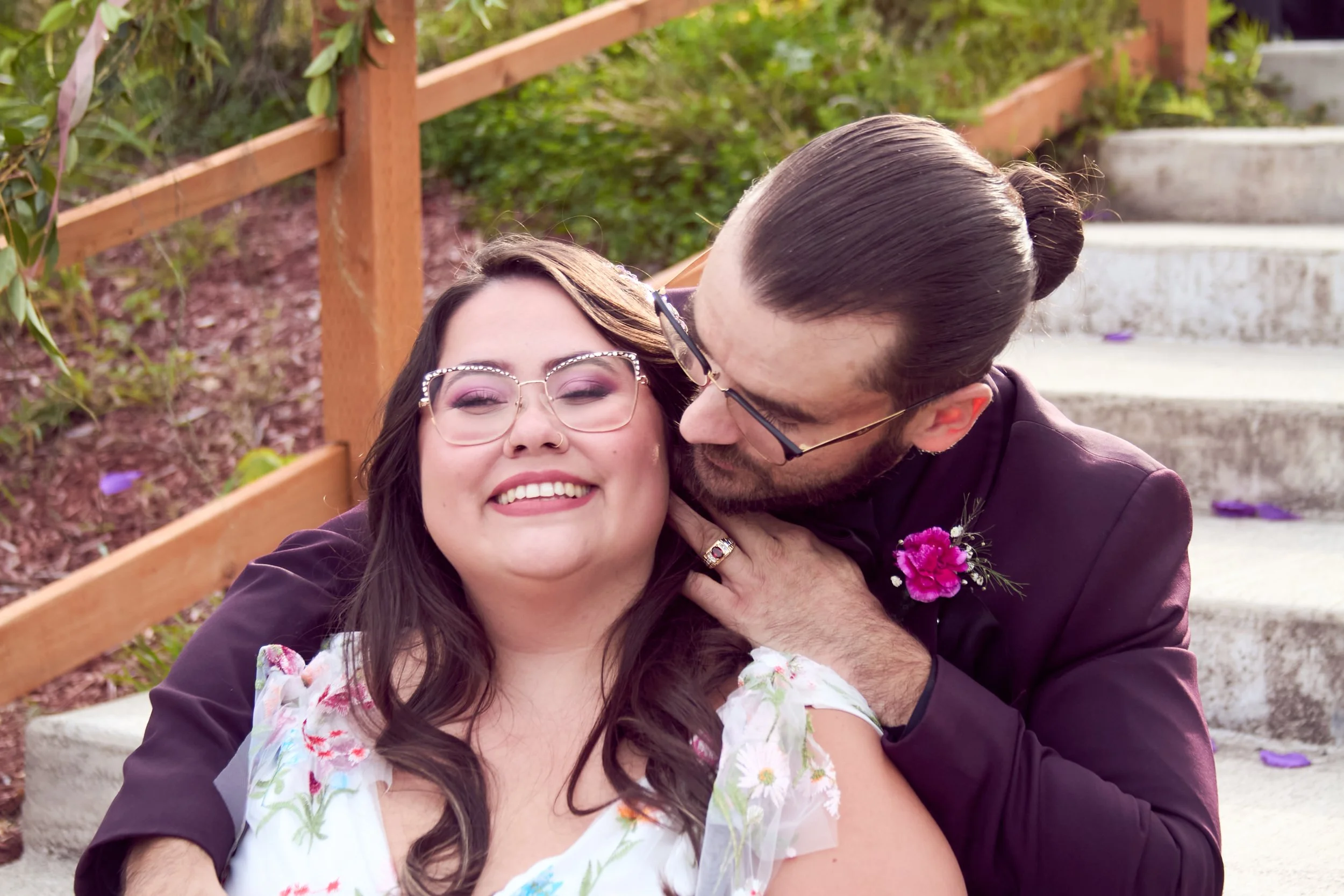 A joyful couple sharing a kiss outdoors on a stone stairway with green bushes and a wooden railing in the background.