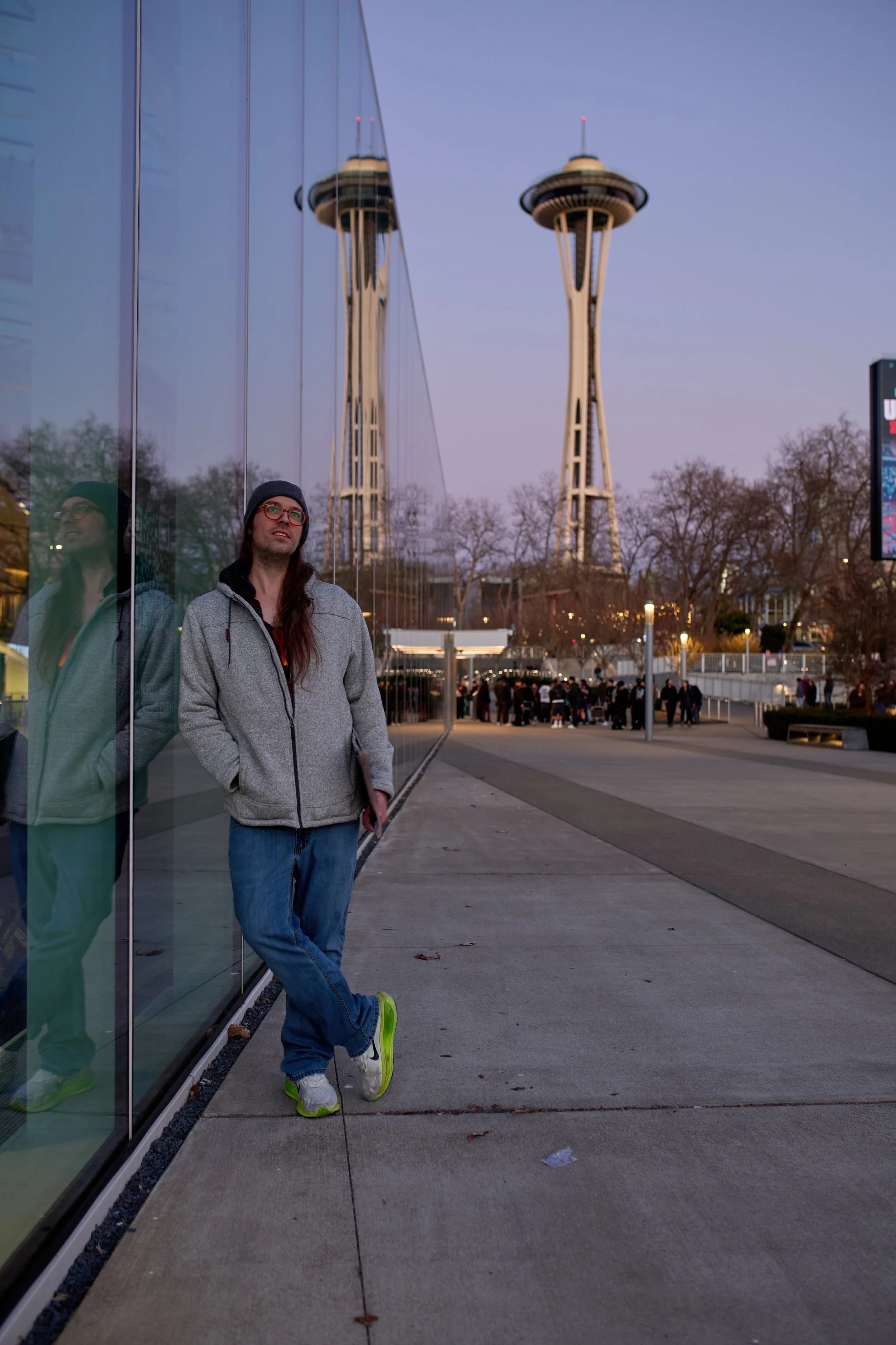 Young man wearing a gray hoodie, jeans, and white sneakers with lime green accents, standing outside near a reflective glass building with the Space Needle in the background, during dusk.