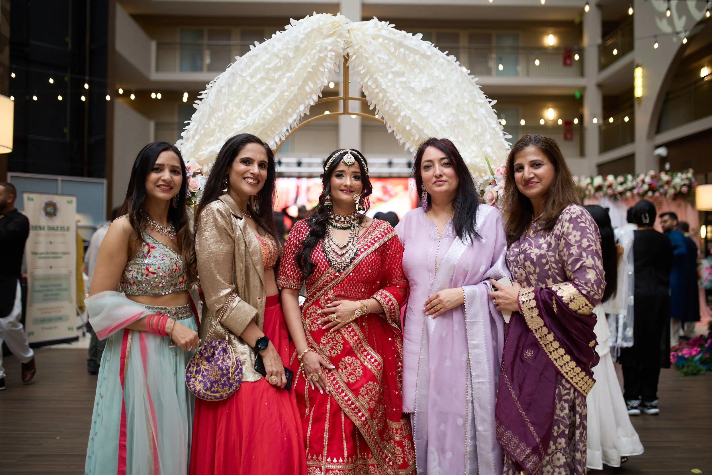 A group of five women dressed in traditional Indian attire at a celebration or wedding event, standing in front of a floral arch with soft lighting.