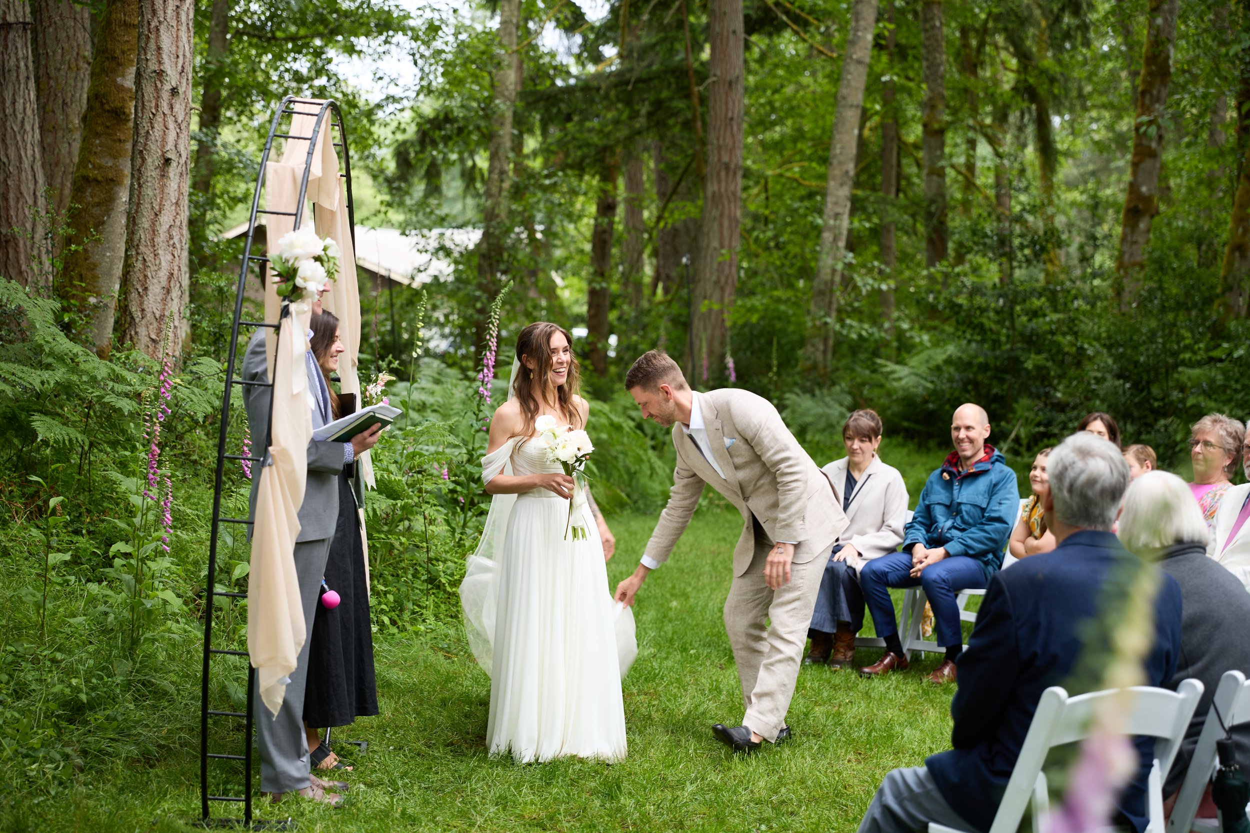 A wedding ceremony taking place outdoors in a wooded area with many guests seated, a bride in a white dress holding flowers, and a groom in a beige suit kneeling before her. The officiant stands nearby, and a decorative outdoor arch with flowers is behind them.
