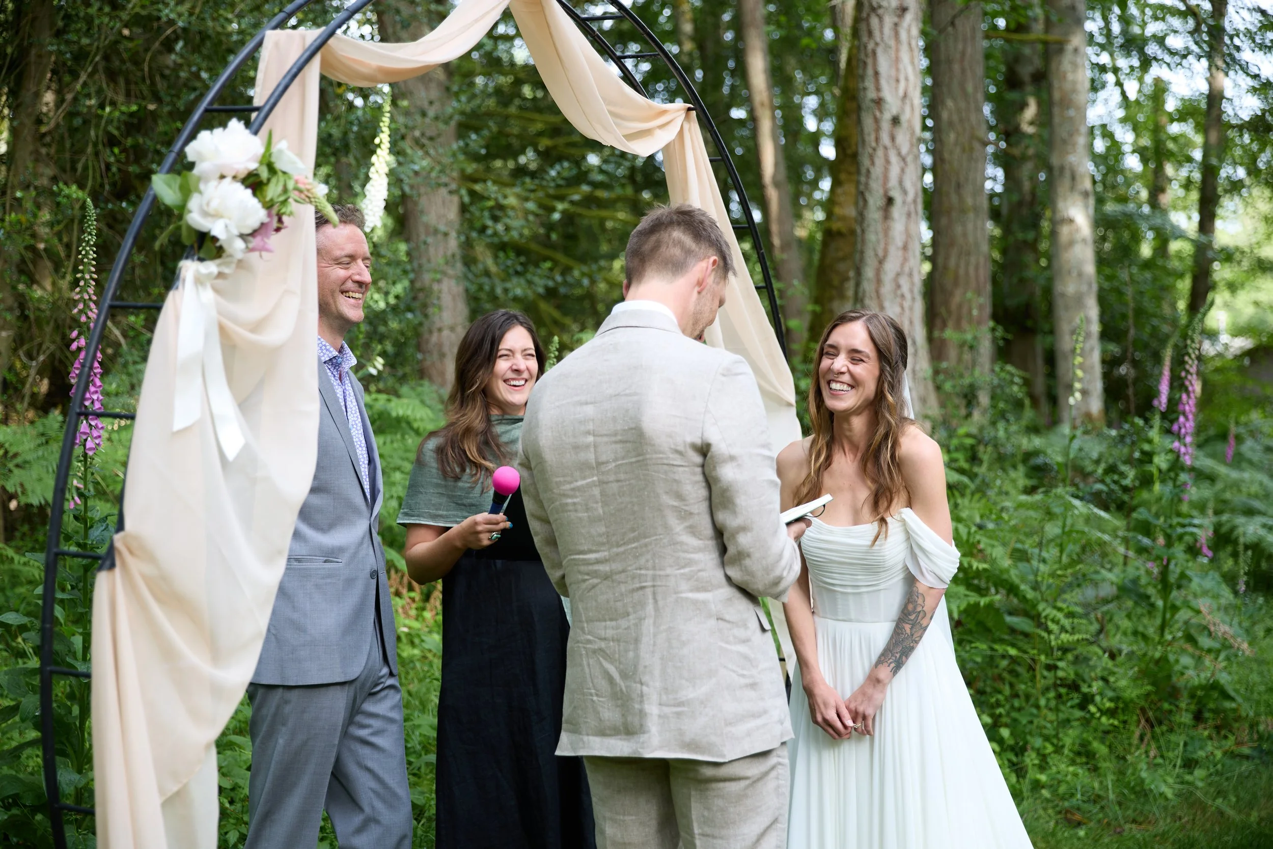 A wedding ceremony in a wooded outdoor setting with five people, including a couple exchanging vows, smiling and holding hands, under a decorated arch with flowers and fabric.