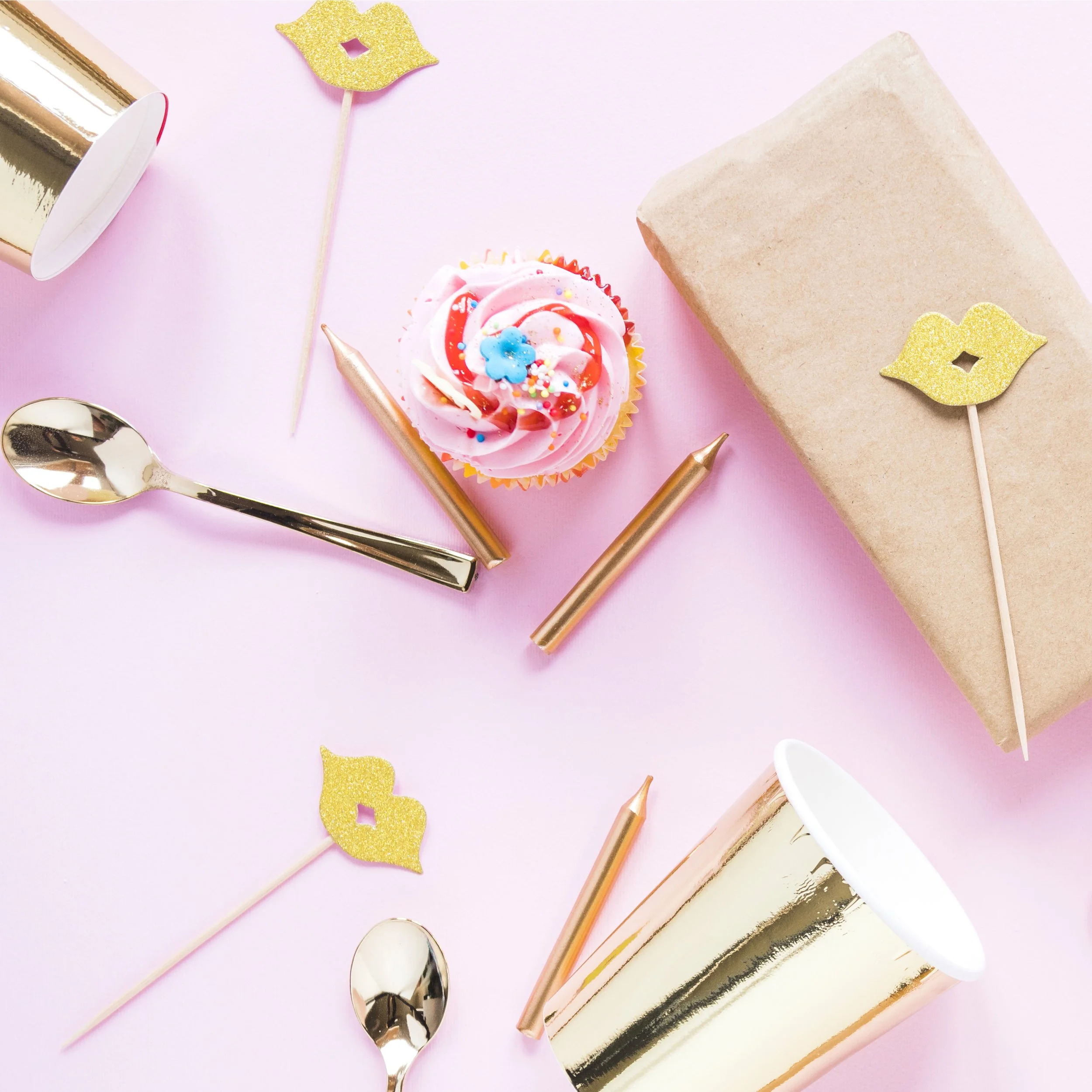 Party table setup with pink cupcake, gold spoons, gold and white cups, gold flags, and a wrapped gift on a pink background.