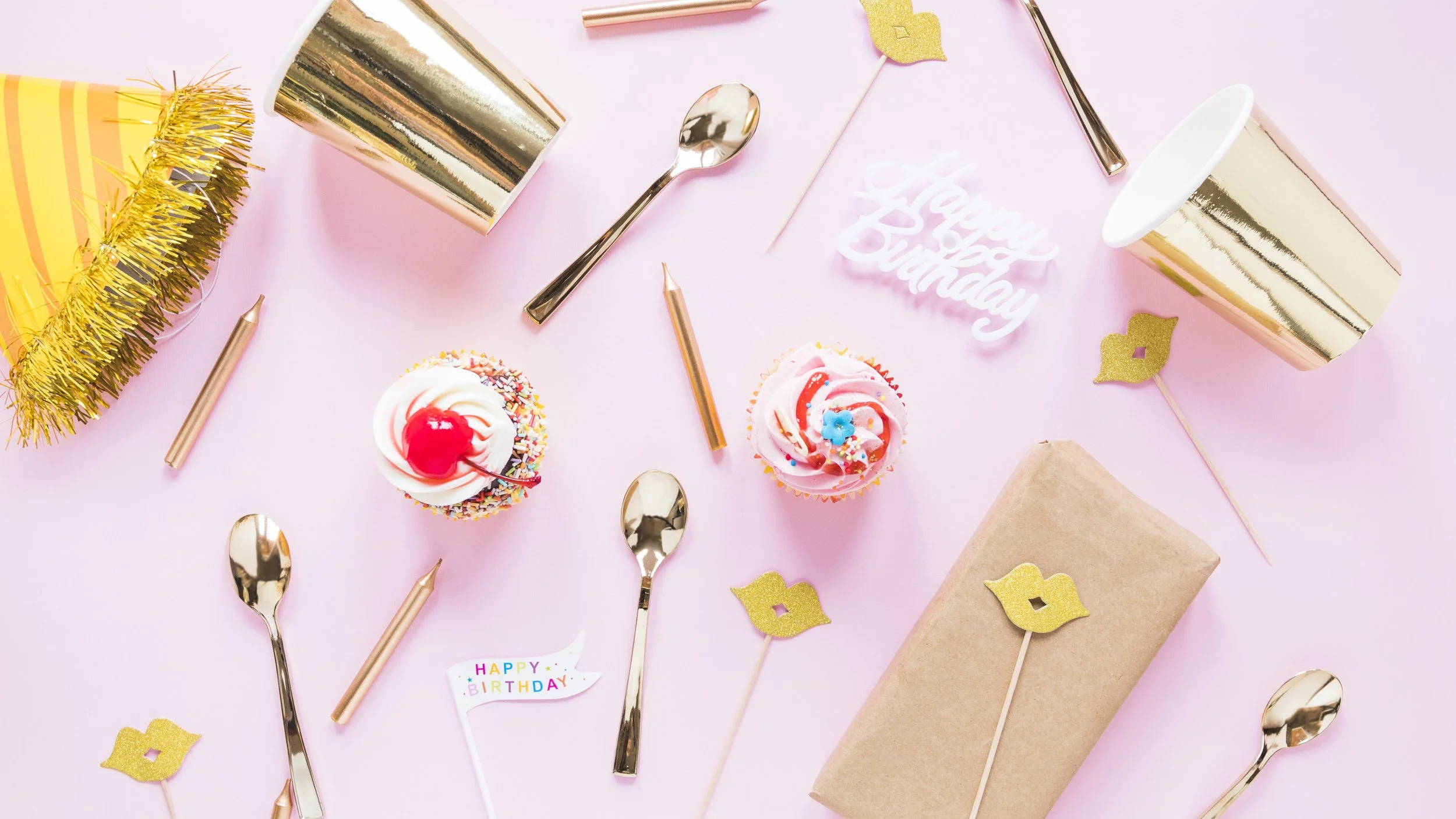 Pink birthday party table with cupcakes, party hats, gold cups, gold spoons, gold and pink candles, gold lips and kiss decorations, a "Happy Birthday" sign, a wrapped gift, and a small birthday flag.