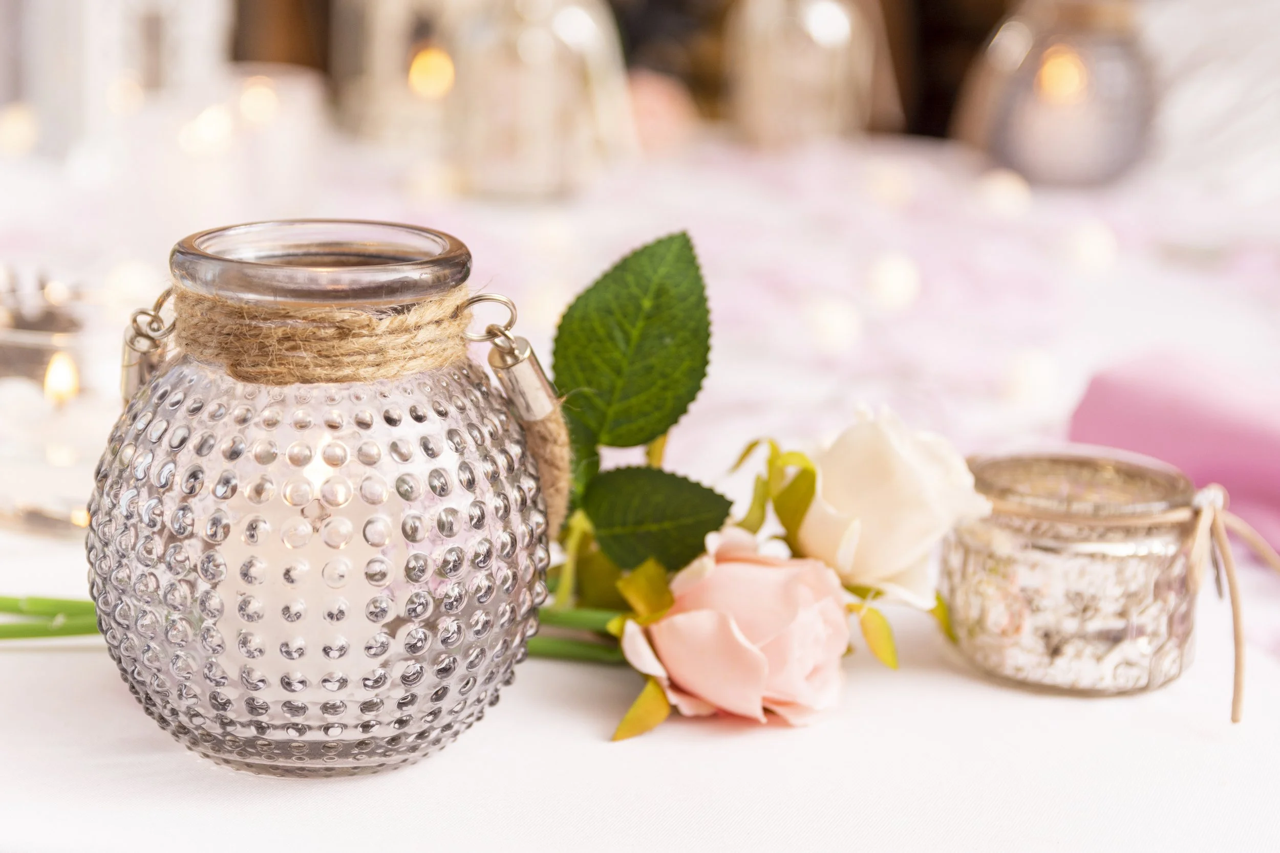 A decorative glass lamp with a textured, dotted surface, wrapped with a jute twine, sitting on a white table. Next to it are pink and white roses with green leaves, and a small jar with a lid, also wrapped with jute. The background is softly blurred 