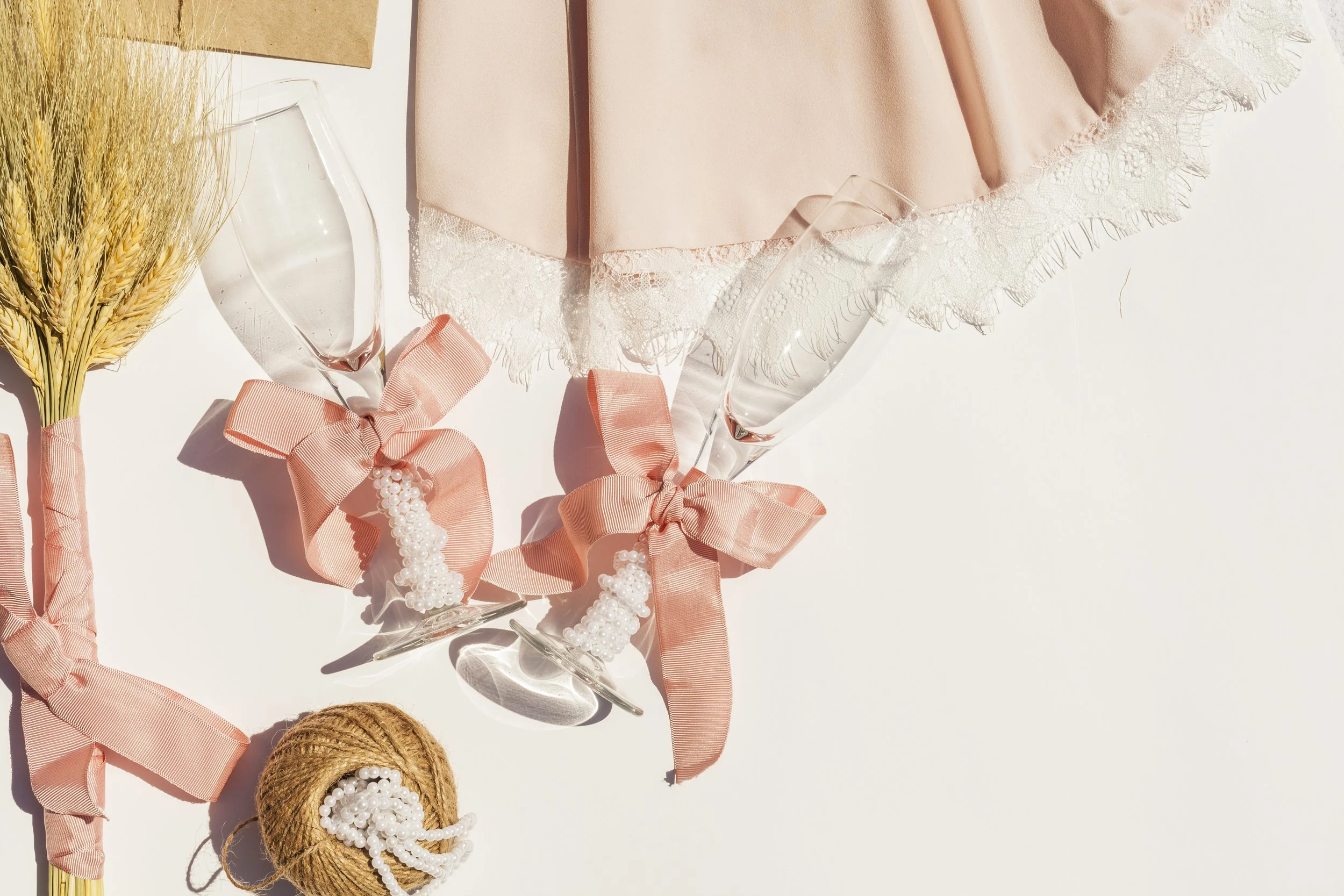 A bouquet of wheat stalks, two champagne flutes with pink bows and pearl decorations, a pink dress with lace trim, and a ball of twine with pearl beads on it placed on a white surface.