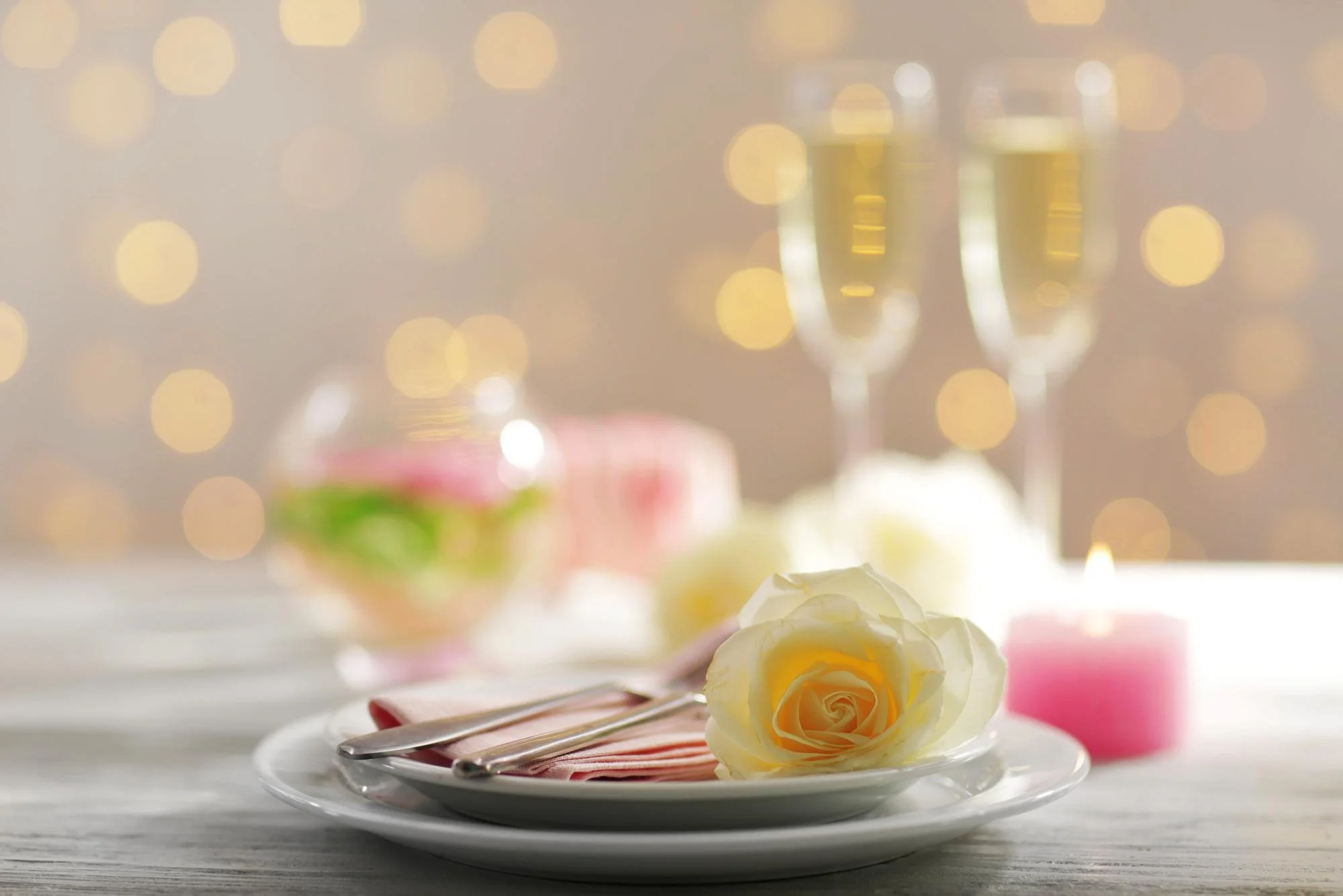 Elegant table setting with a white rose, pink napkin, and silverware on a plate, with champagne glasses and blurred lights in the background.