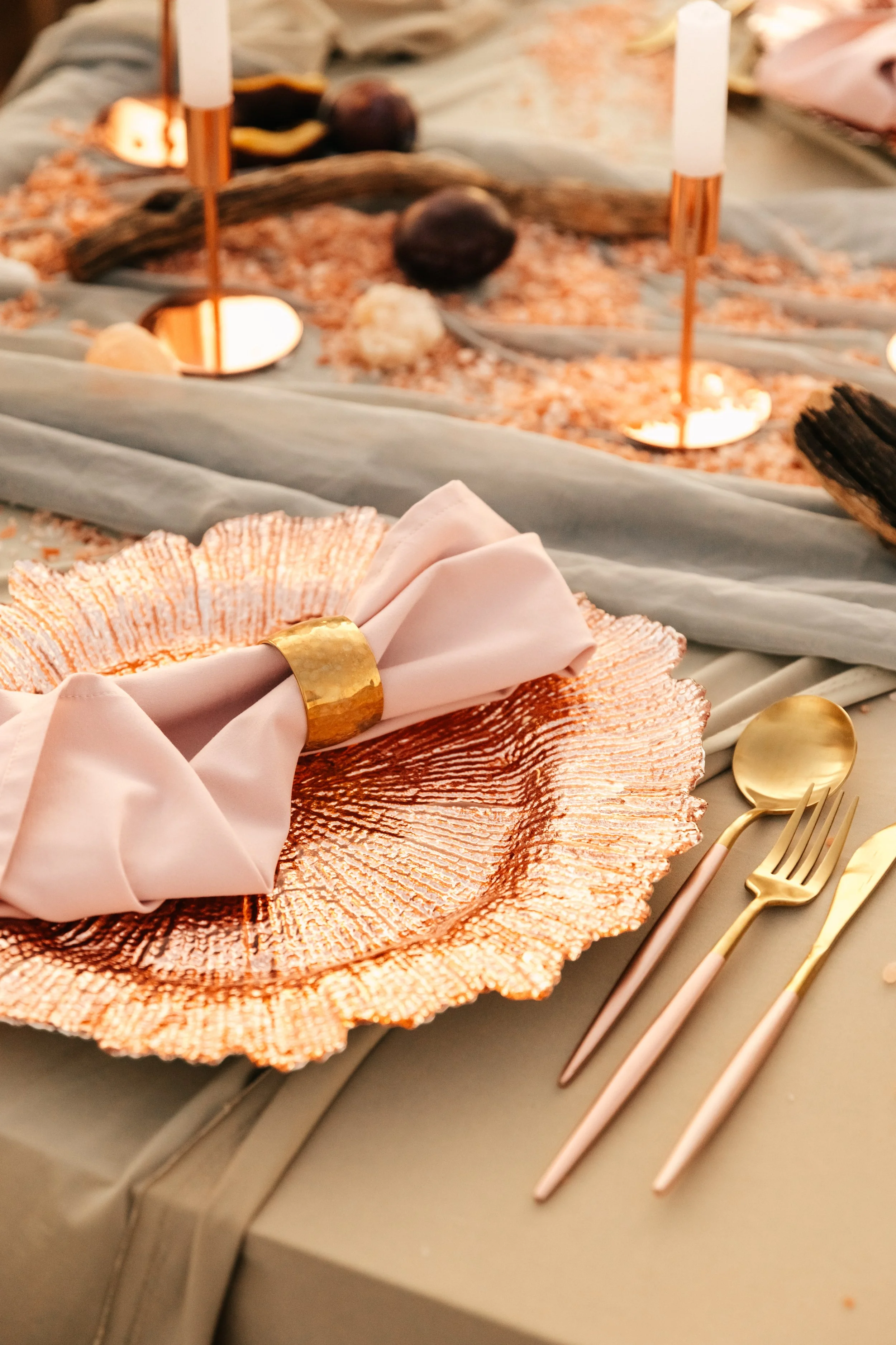 Elegant table setting with a rose gold decorative plate, gold cutlery, and a neatly folded light pink napkin secured with a gold napkin ring, on a table with a draped cloth and a decorated centerpiece in the background.