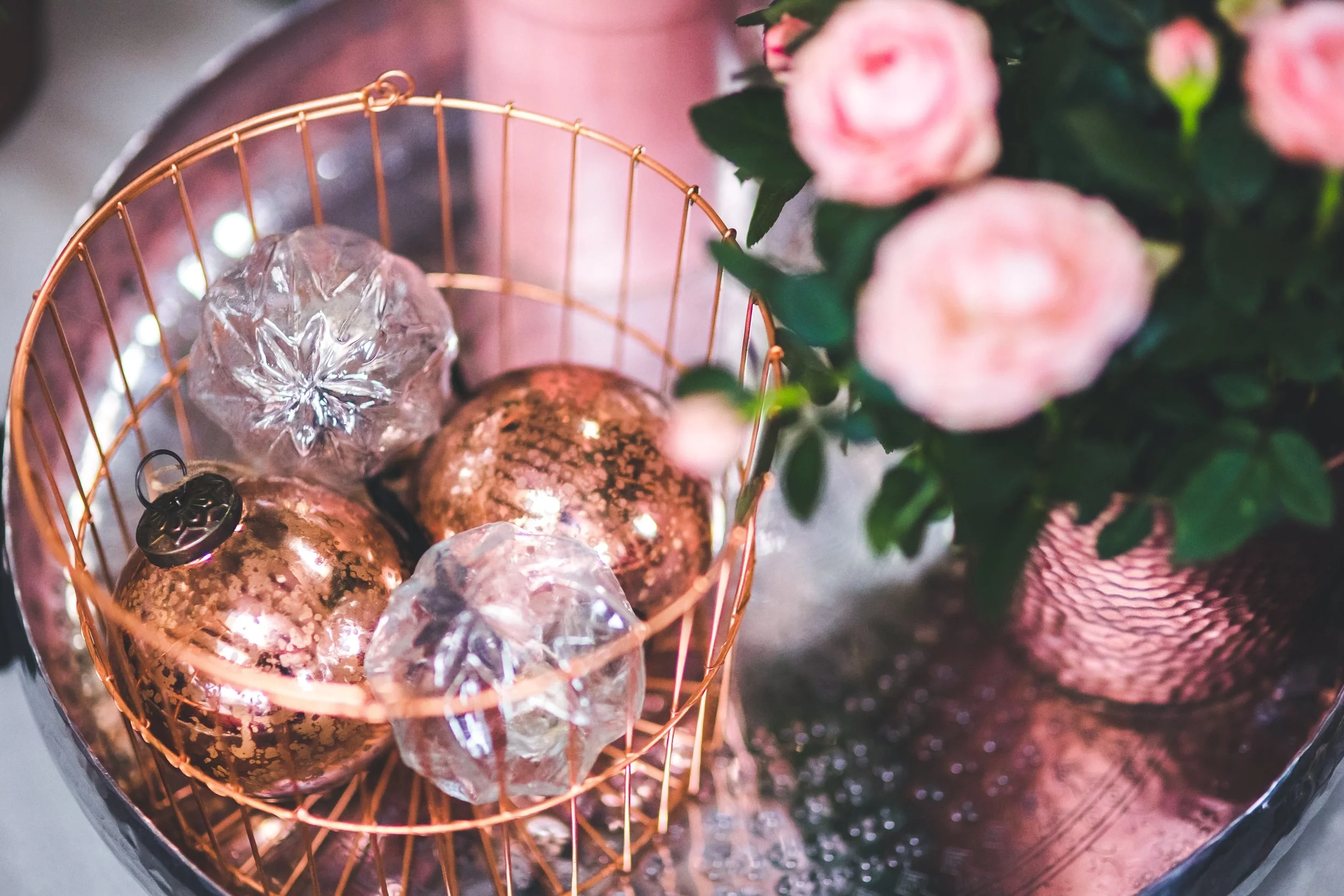 A rose gold wire basket holding Christmas ornaments, some gold and others clear with a reflective surface, next to a pot with pink roses in a pink vase, all on a reflective surface.