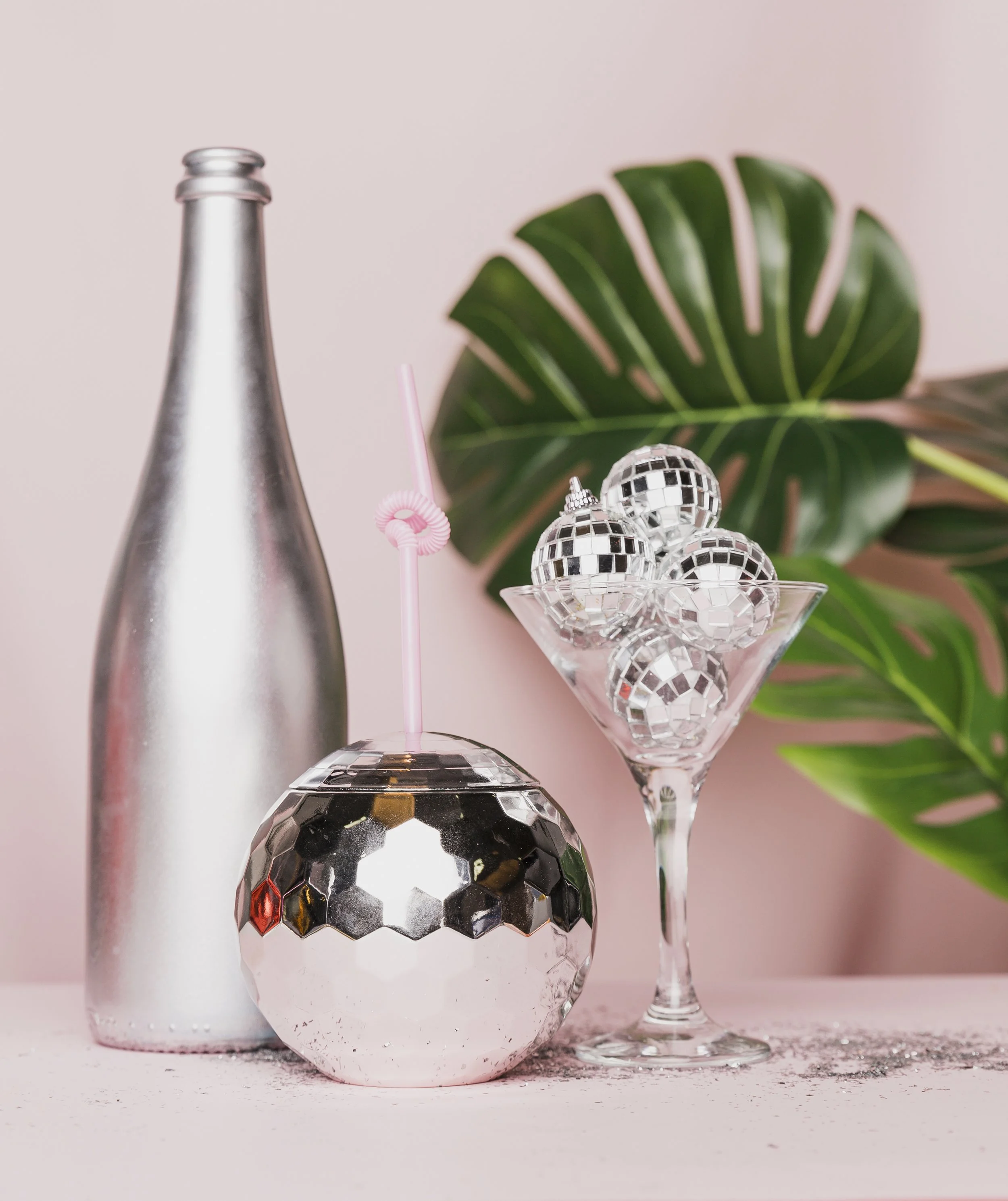 Silver champagne bottle, a mirrored disco ball with a pink straw, and a martini glass filled with small silver disco balls on a pink surface with green tropical leaves in the background.