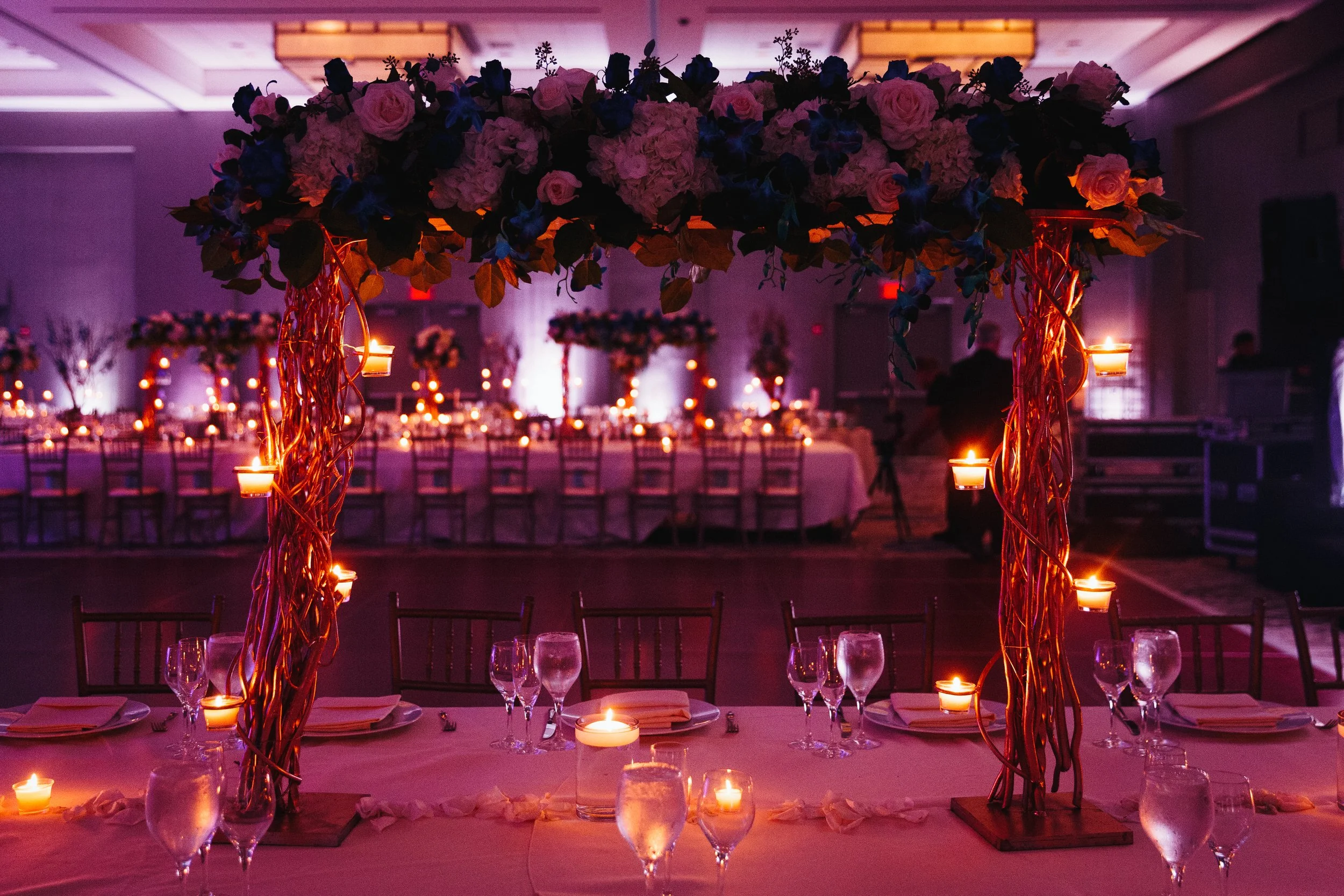 Elegant banquet hall decorated with tall floral centerpieces, featuring pink roses and blue flowers, surrounded by lit candles and set tables with glassware and napkins for a formal event or wedding reception.