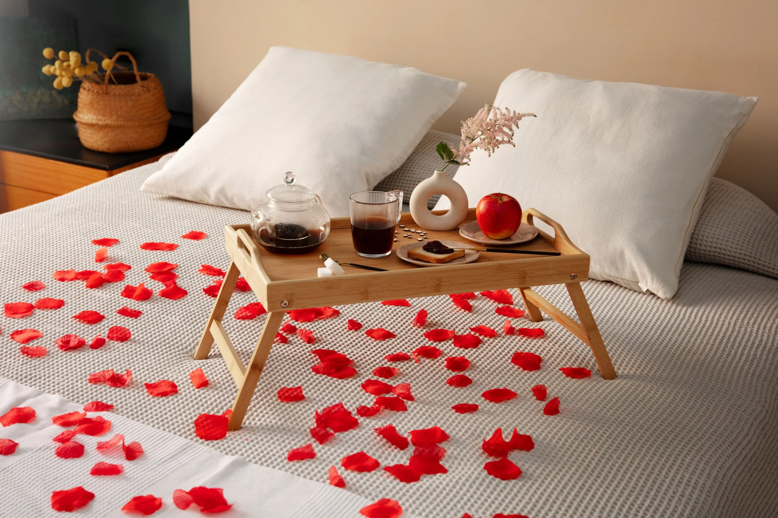 A bedroom with white pillows on a bed covered in rose petals, a wooden tray holding a teapot, glass of tea, plate with toast and jam, a red apple, and a small vase with pink flowers.