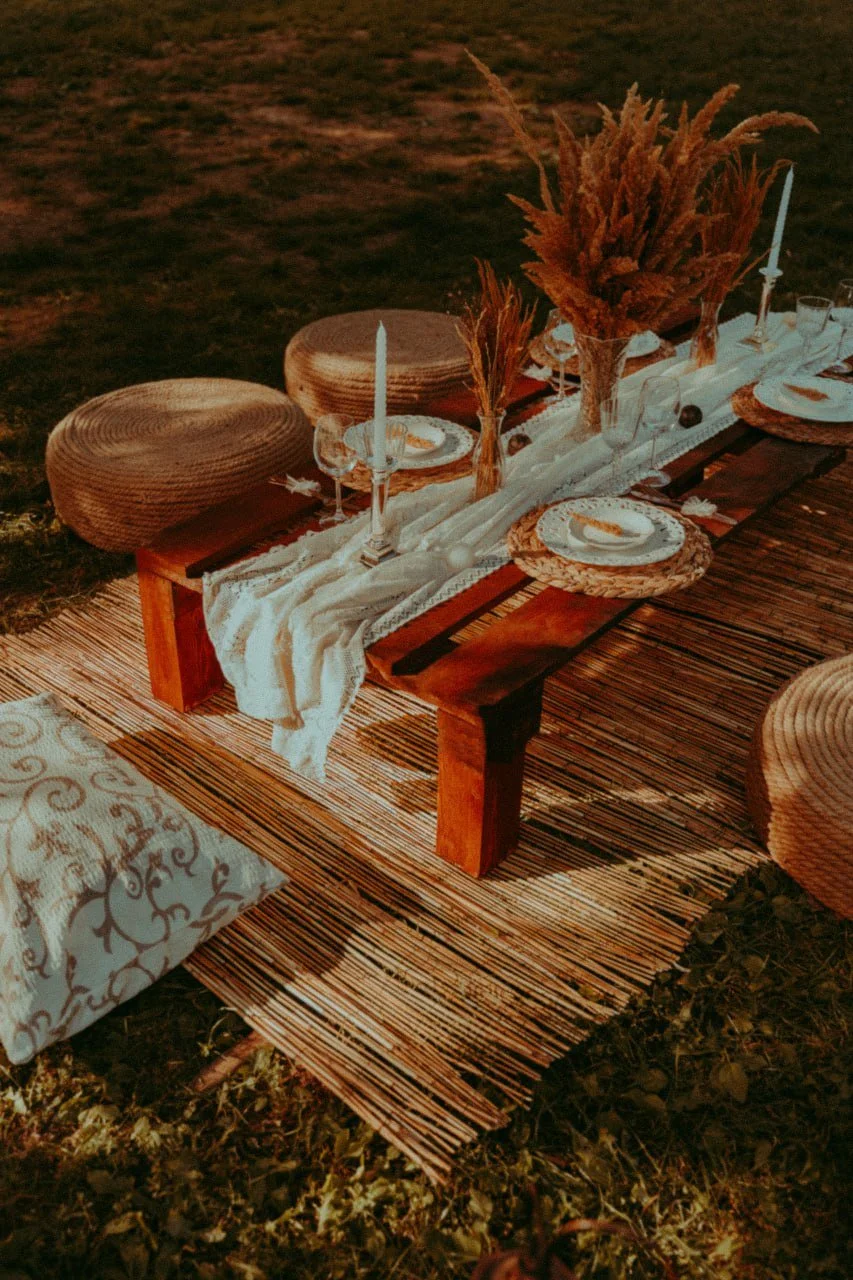 A rustic outdoor dinner table setup with wooden benches, woven round seats, and a striped table runner. The table is decorated with flowers, candles, and dinnerware for an elegant gathering.