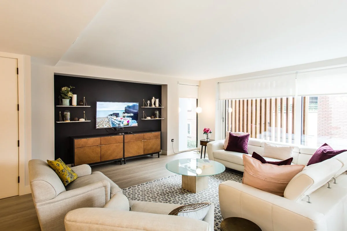 Spacious and relaxing living space with cream Italian corner suite, burgundy and pink cushions, round glass coffee table, large wall hung TV and wall to ceiling windows with roller blinds.