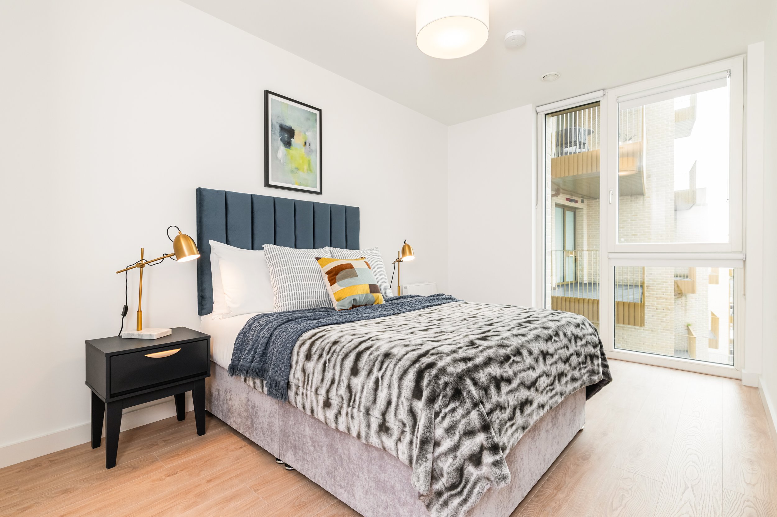 Minimalist bedroom with white walls, large windows, a blue upholstered headboard, bed with assorted pillows, grey throw blanket, and a black nightstand with gold lamps.