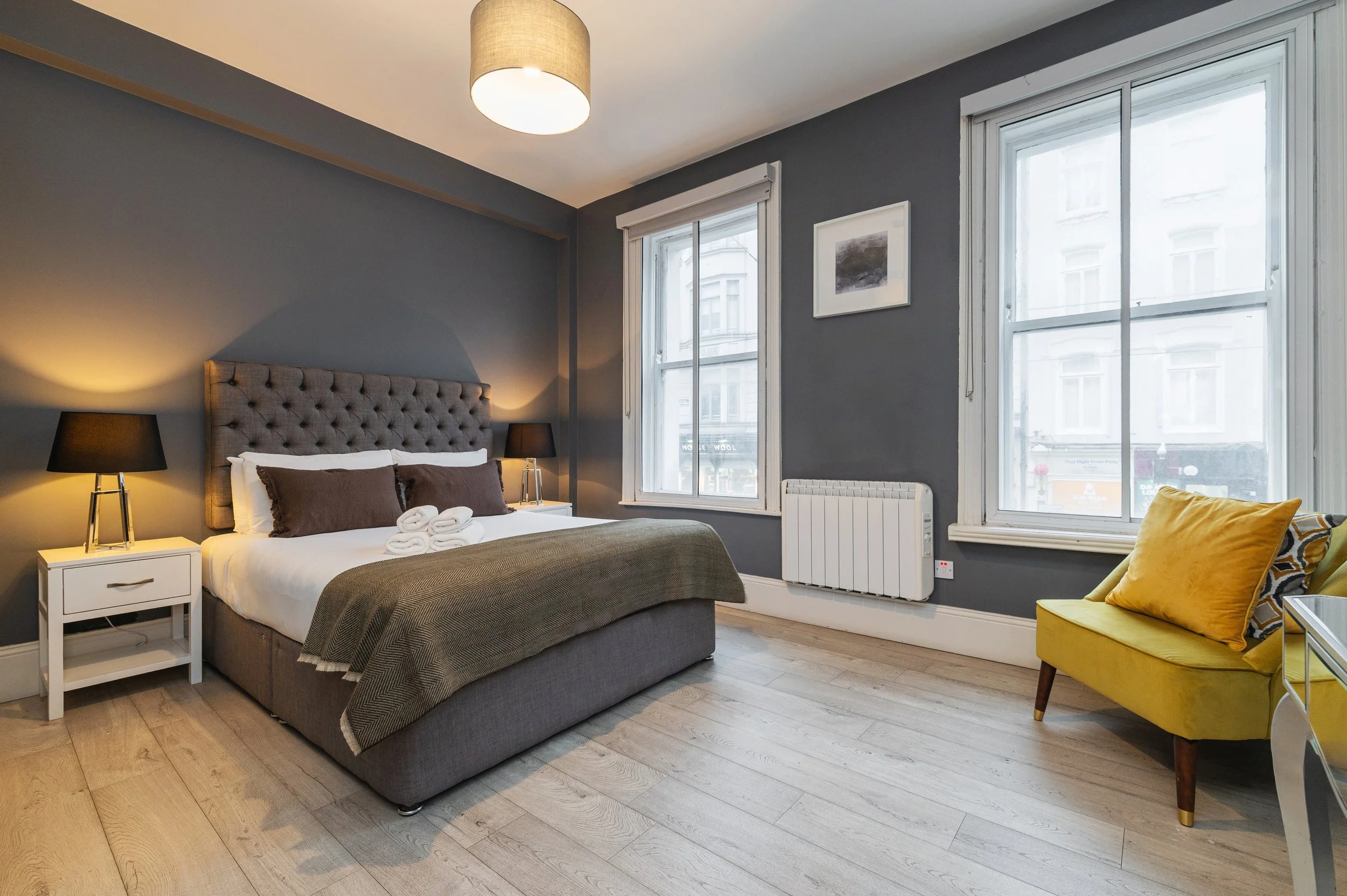 A modern bedroom with a king size bed, gray upholstered headboard, white linens, and pillows. Nightstands with black lamps on either side. Two windows with white blinds and a small yellow chair in the corner.