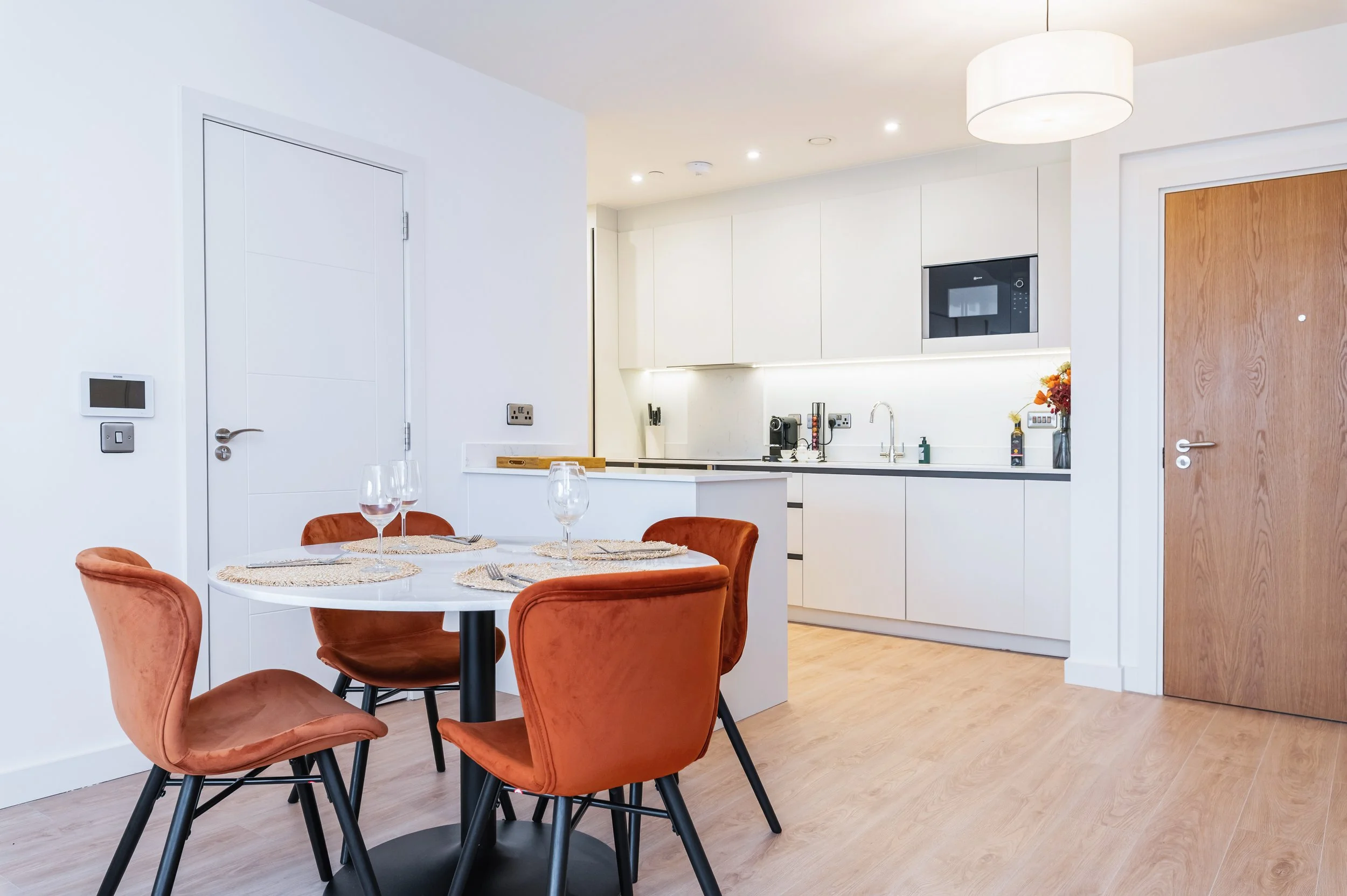 Modern white kitchen with wooden dining table and brown velvet chairs, built-in appliances, and minimal decor.