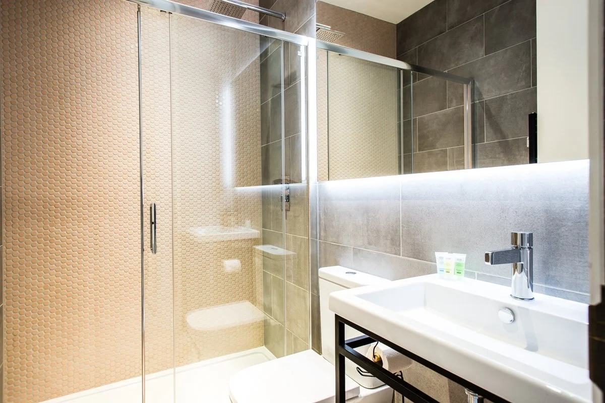 Pale pink mosaic shower room with sliding shower door, wall hung toilet, large white ceramic sink with black metal stand and chrome tap.