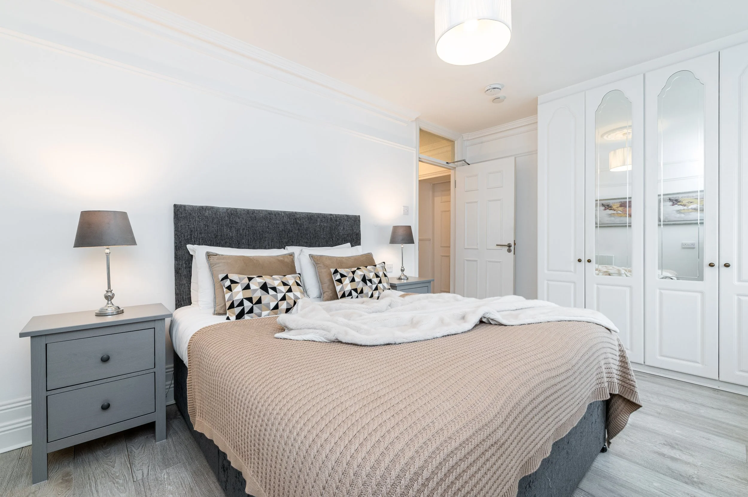 A bedroom with a bed featuring a gray headboard, white bedding, and decorative pillows, a cream and taupe throw blanket, grey nightstands with lamps, a white wardrobe with mirrored doors, and a hanging ceiling light.