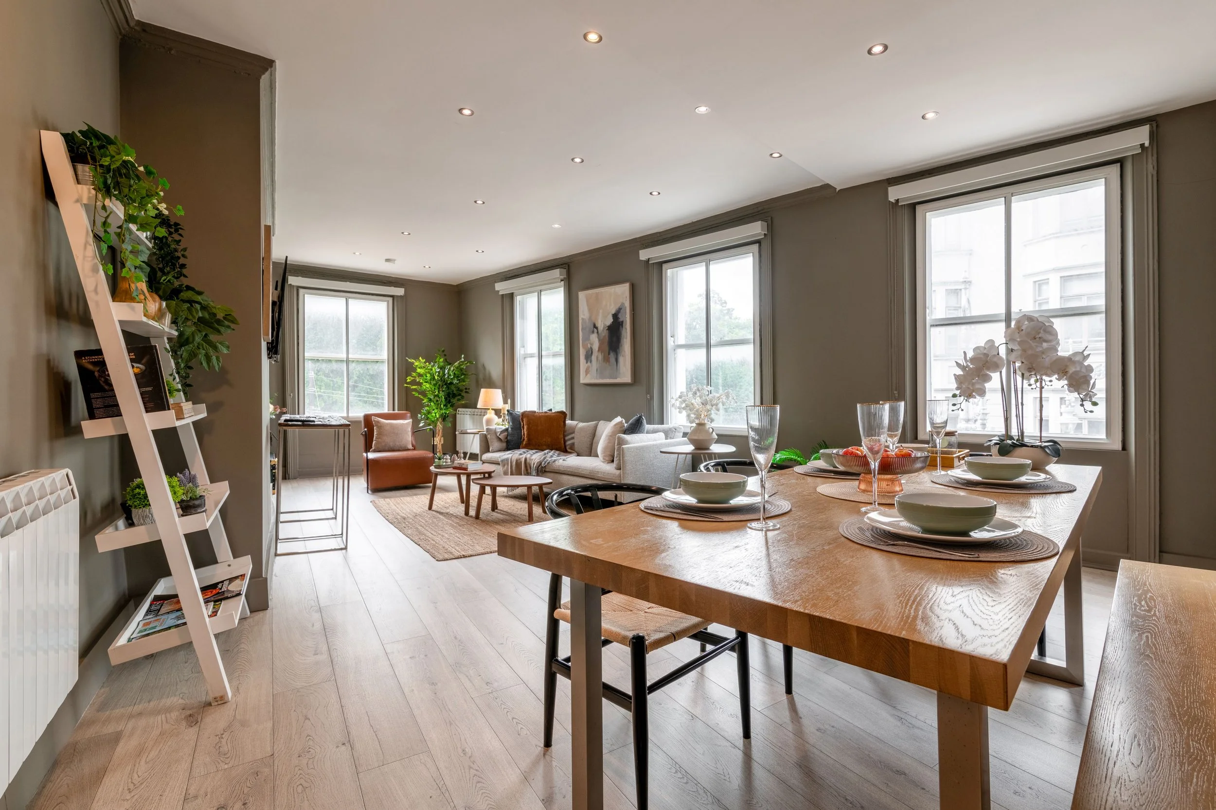 Living room with mink walls, modern furniture including a cream sofa, brown leather armchair, a wooden console table, a flat-screen TV, a wooden coffee tables, and large windows with vertical blinds. There is a large dining table with a seating bench