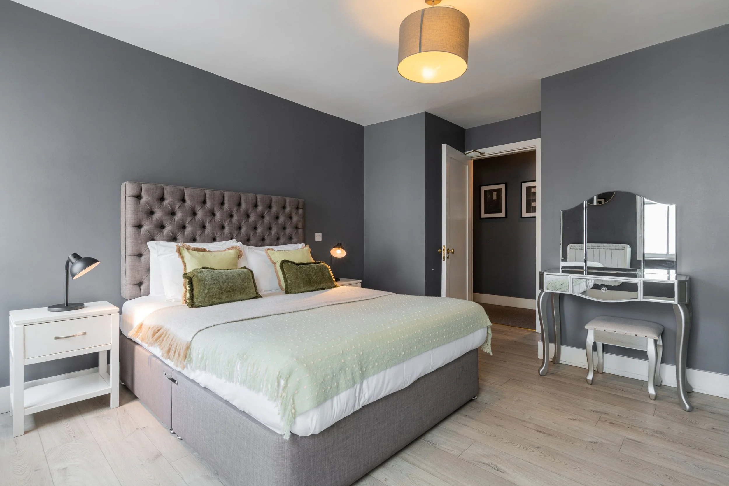 Modern bedroom with gray walls, a large bed with gray headboard, white bedding, and green cushions and throws. There is a white nightstand with black lamps on each side of the bed. A mirrored vanity desk with a padded stool is by the wall with a yell