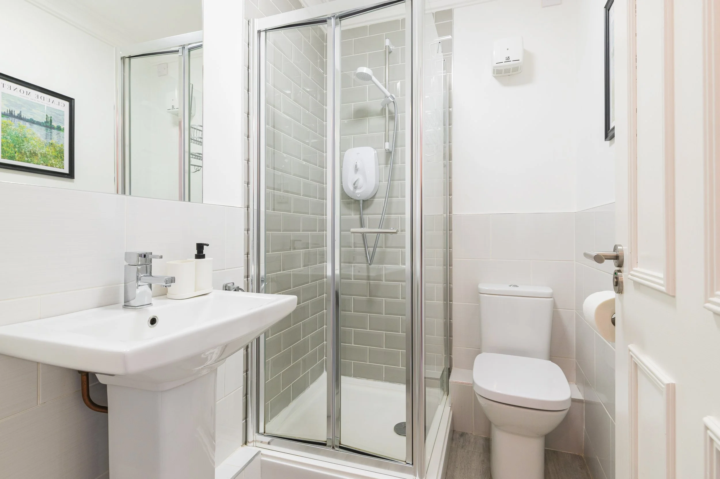 Small modern ensuite shower room with a corner shower, white toilet, and white sink, decorated with framed artwork and minimal accessories.