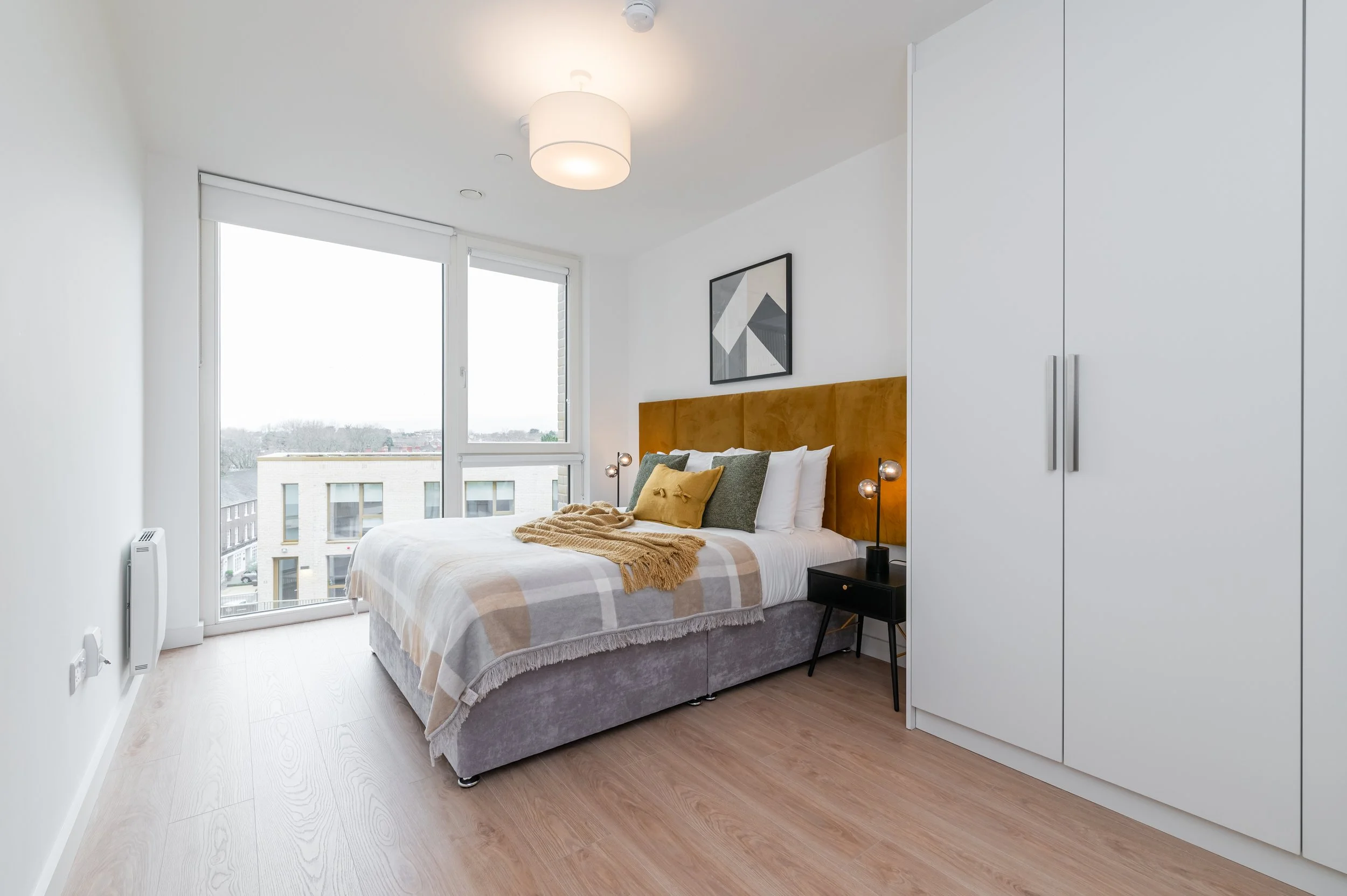 Bright, modern bedroom with a bed featuring a mustard headboard, white bedding, and patterned pillows. Next to the bed is a black nightstand with a modern double lamp. A large window with blinds. The room has light wood flooring, white walls, and a c