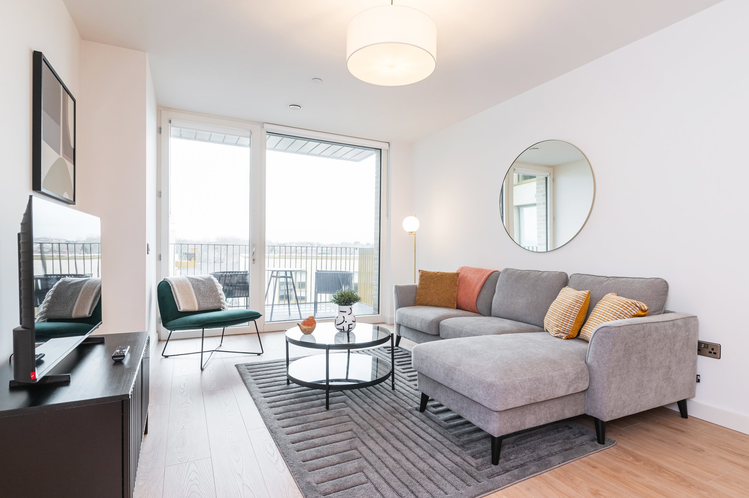 Living room with beige sofa, glass coffee table, TV stand, and balcony with outdoor seating overlooking trees.