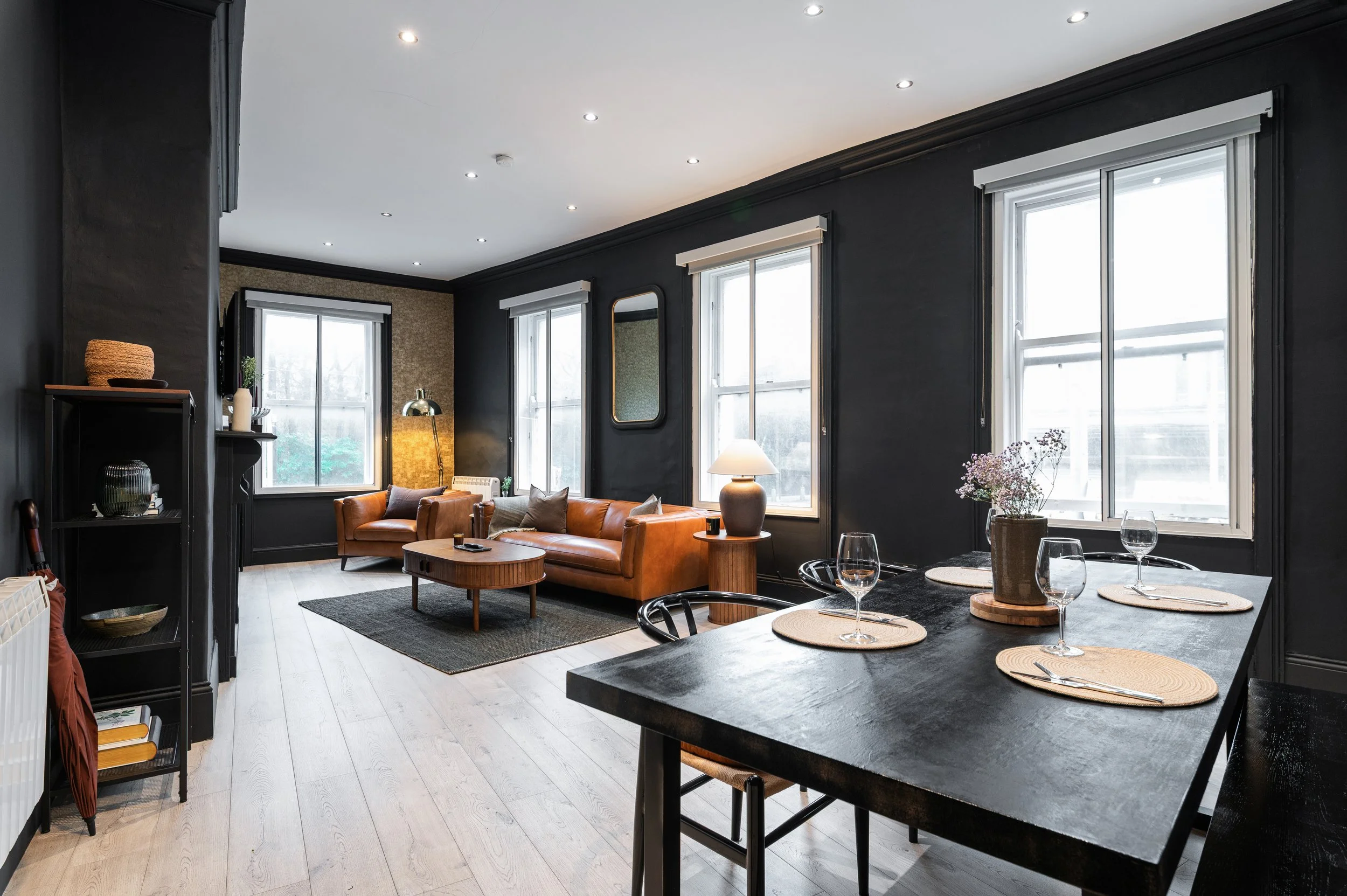 A living room with a brown leather couch, a wooden coffee table and matching side table, a decorative fireplace, a black ladder shelf with decor, large windows, and a dark rug on light wood flooring. A large 8 seater dining table with a bench and cha