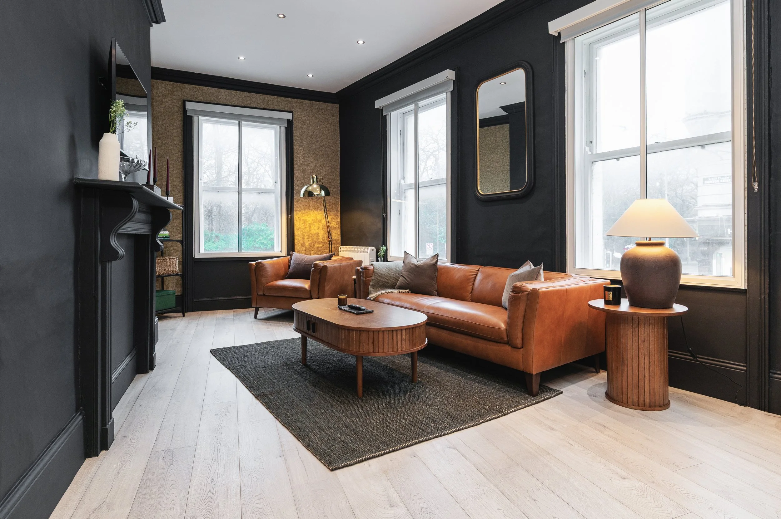 Living room with dark grey walls and large windows. Modern furniture includes a brown leather sofa, a brown wooden coffee table and matching side table and a gold floor lamp. A rectangular mirror hangs above the couch. A fireplace with decorative mol