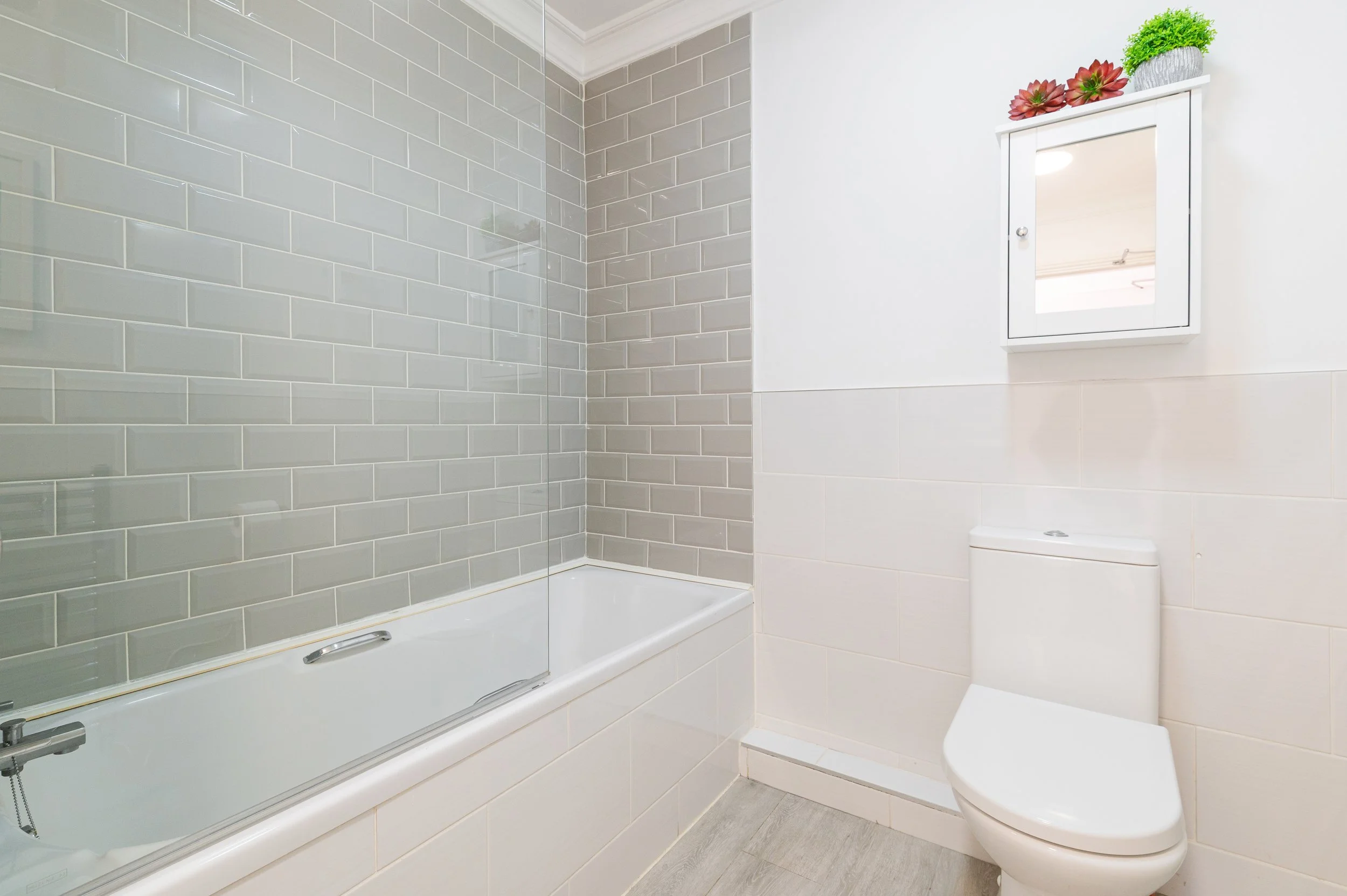A small, modern bathroom with a white toilet, shower over bath, a wooden wall hung cabinet with decorative plants. The room has light-colored walls and wood-look flooring.