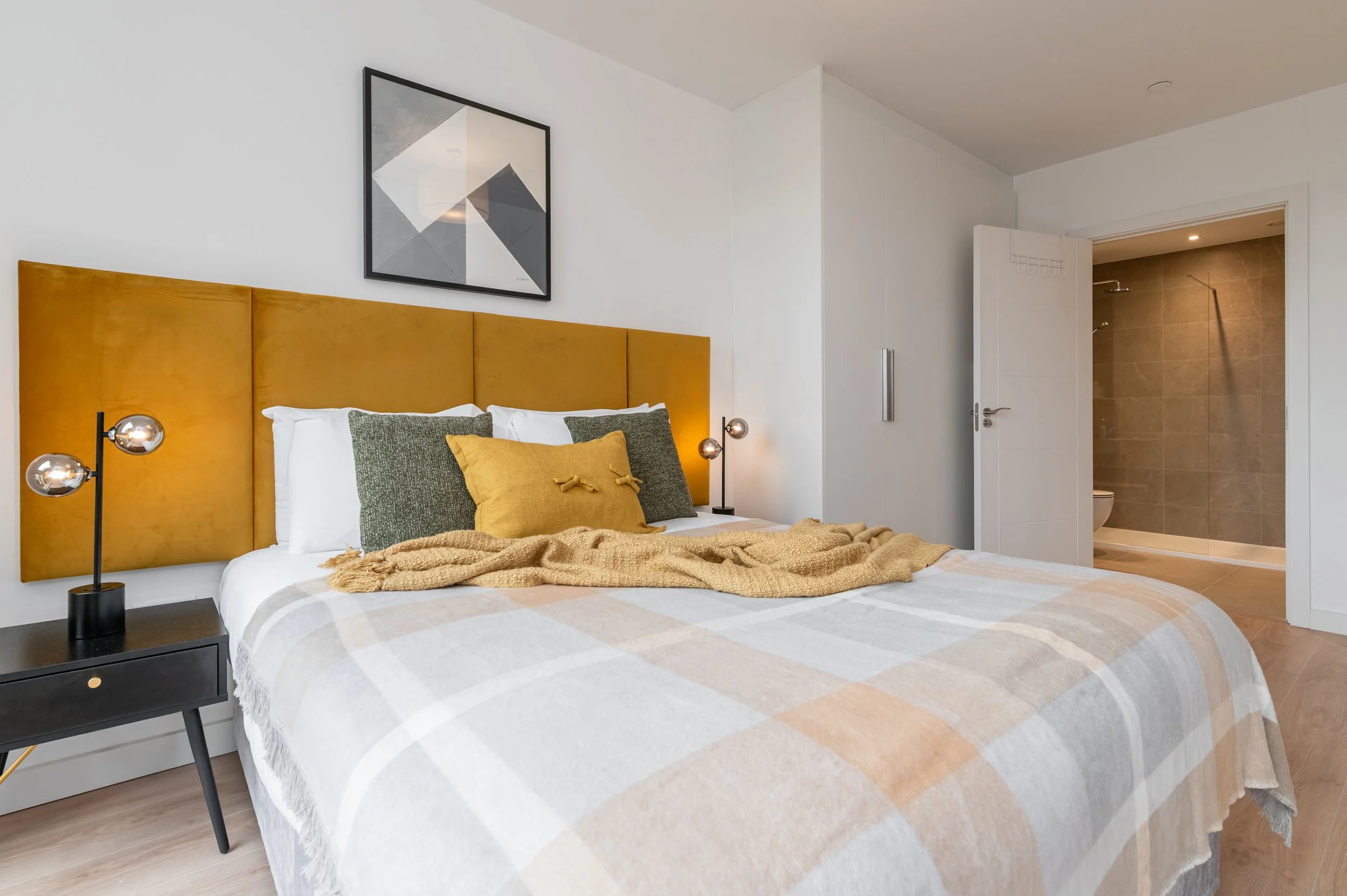 Bright, modern bedroom with a bed featuring a mustard headboard, white bedding, and two pillows. The room has light wood flooring, white walls, and a ceiling light fixture.