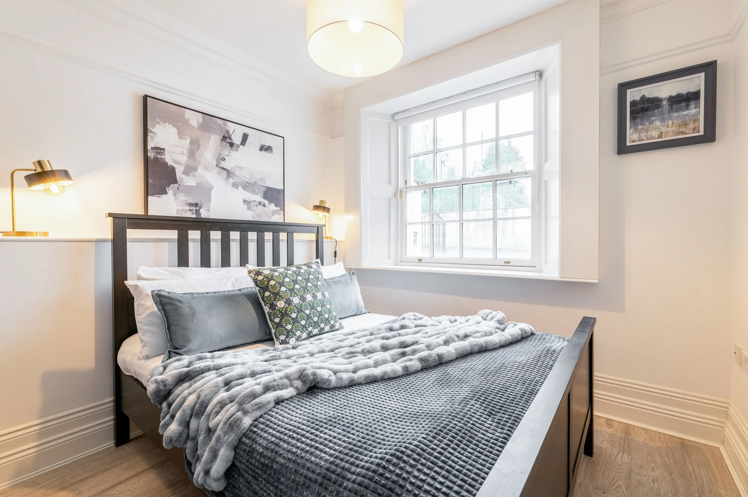 A bedroom with a double bed featuring a wooden bedframe, white bedding, and decorative pillows, a navy throw blanket, grey artwork and a sash window.