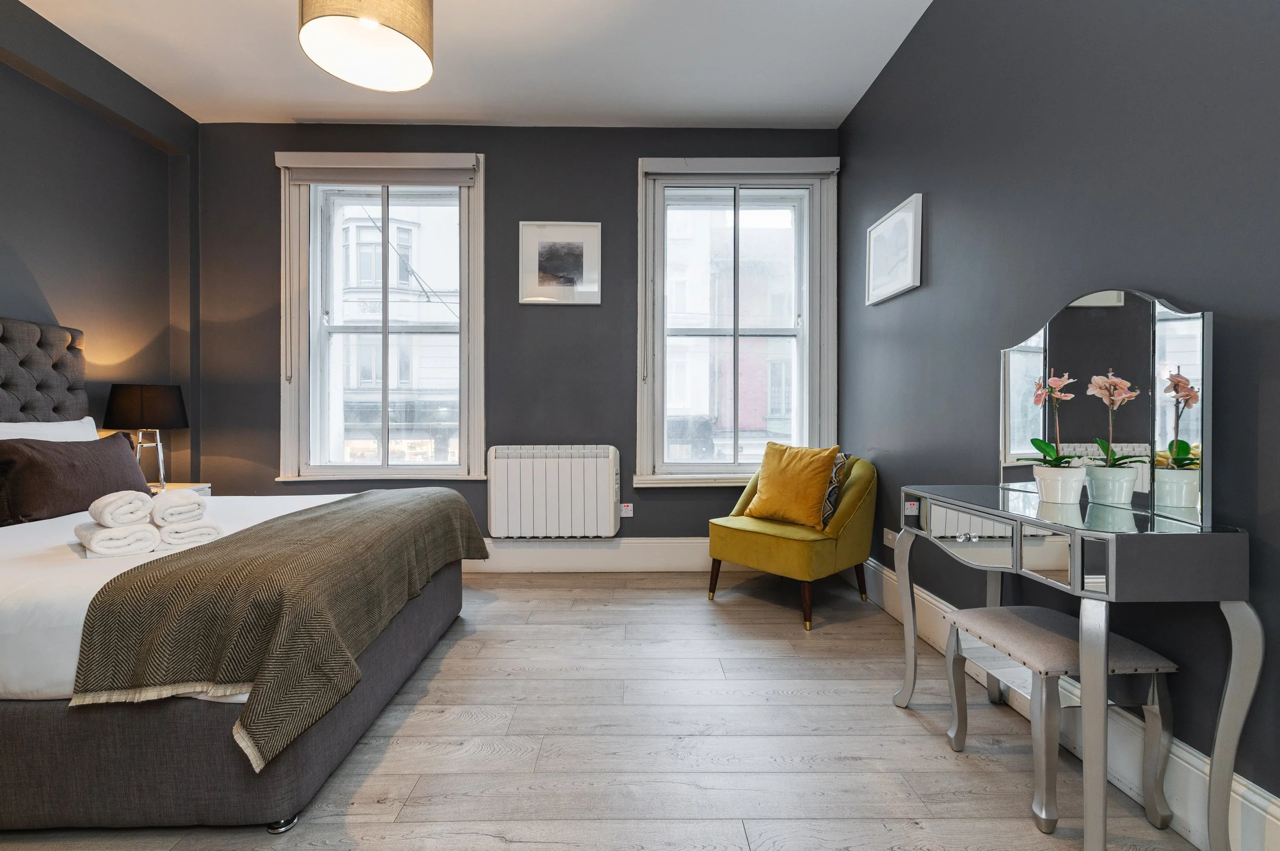 Modern bedroom with gray walls, a large bed with gray headboard, white bedding, and dark cushions. There is a white nightstand with black lamps on each side of the bed. A mirrored vanity desk with a padded stool is by the wall with a yellow armchair 