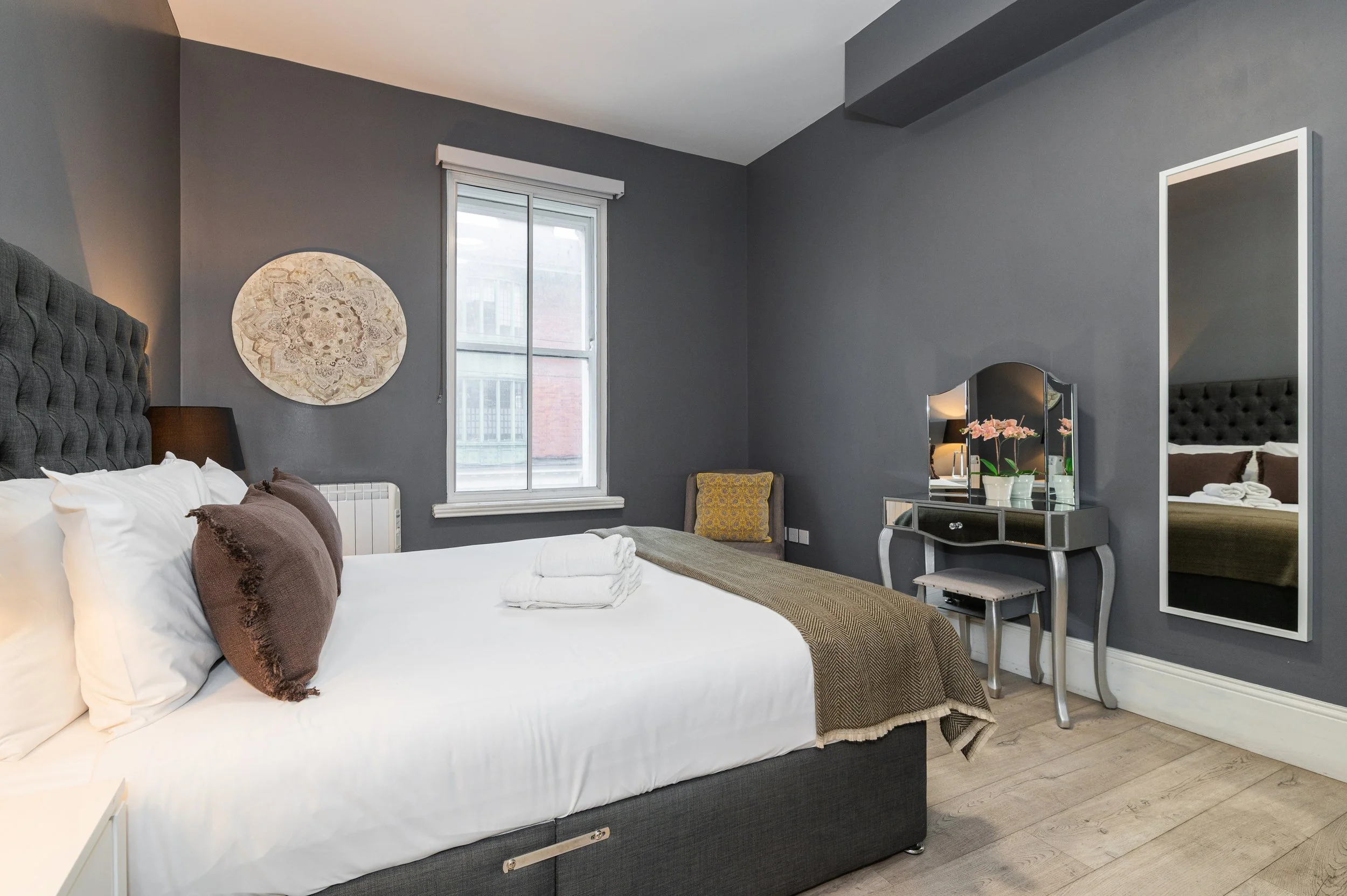 A modern bedroom with a king size bed, gray upholstered headboard, white linens, and pillows. One window with white roller blinds and a small chair in the corner. A mirrored vanity table with padded seat.
