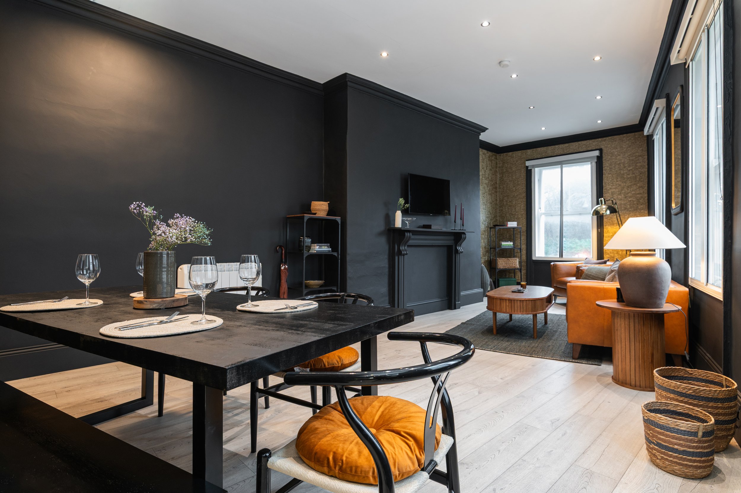 Living room with dark walls, modern furniture including a brown leather sofa, wooden chairs, a fireplace, a flat-screen TV, a wooden coffee table, and large windows with vertical blinds. There is a large dining table with a seating bench and chairs.
