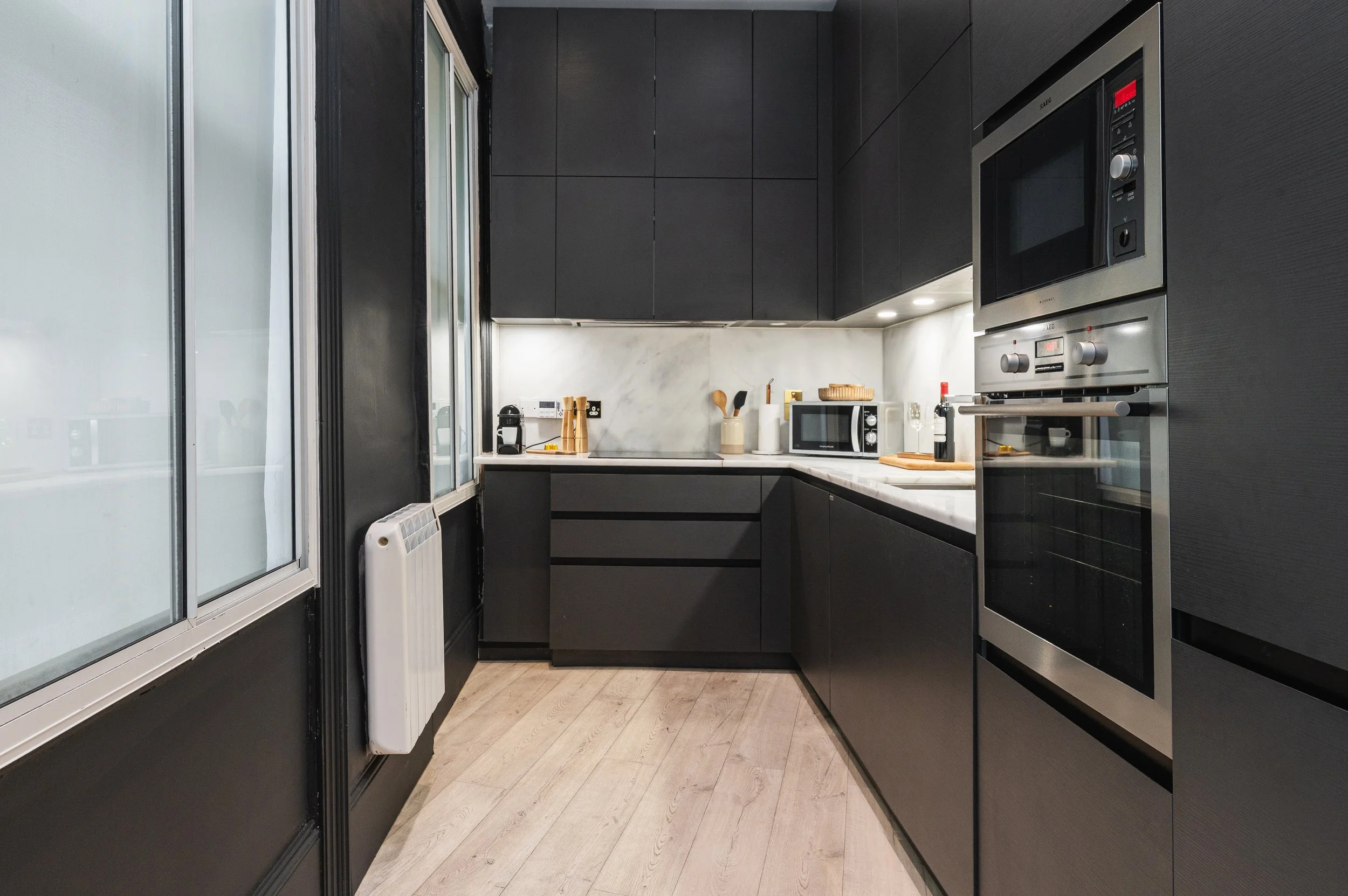 Modern kitchen with black cabinets, white marble countertops and backsplash, stainless steel appliances including toaster, microwave, electric kettle, and sink with chrome faucet.