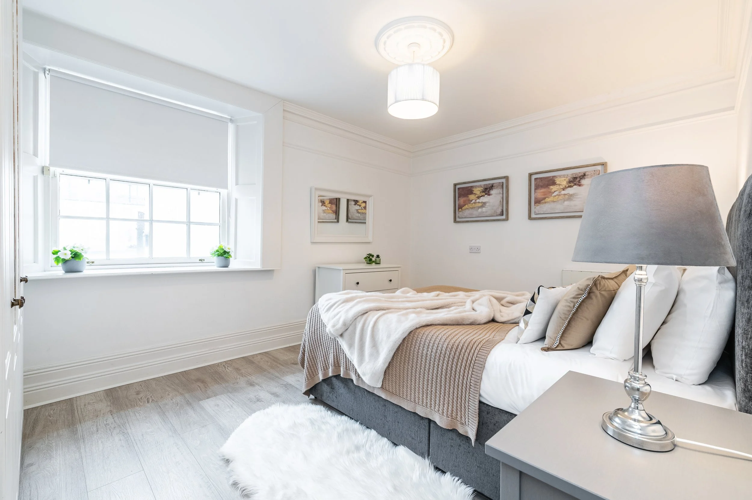 A bedroom with a bed featuring a gray headboard, white bedding, and decorative pillows, a cream and taupe throw blanket, grey nightstands with lamps, a white wardrobe with mirrored doors, and a hanging ceiling light.