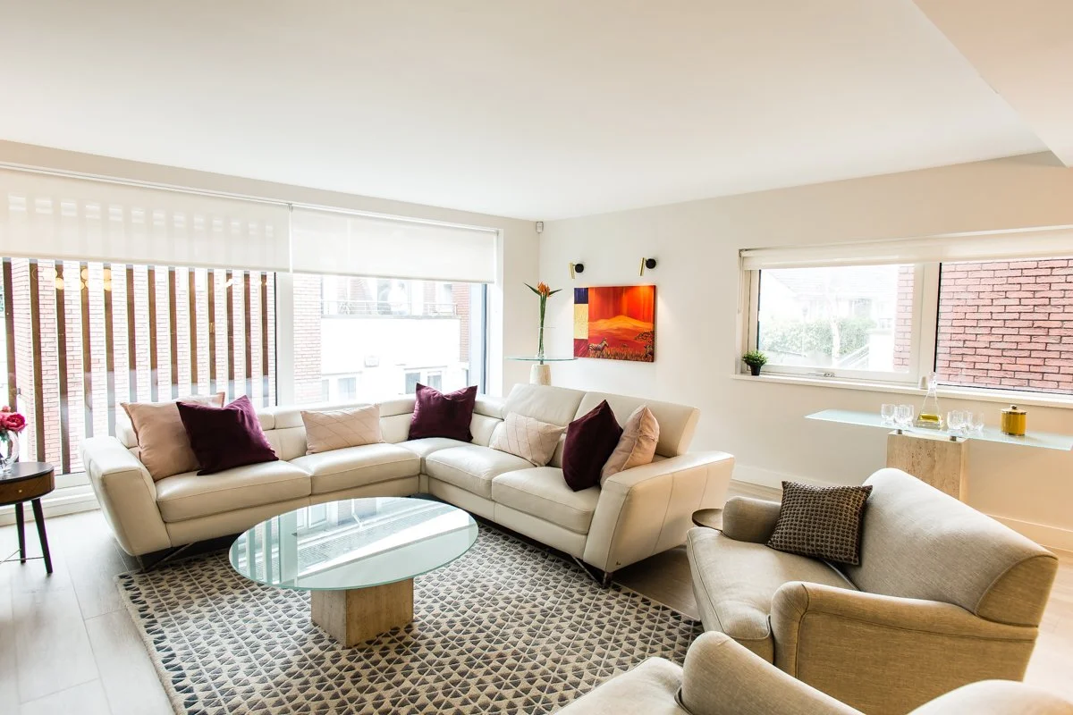 Beautiful living space with cream Italian corner suite, burgundy and pink cushions, round glass coffee table, red artwork with overhead lighting.