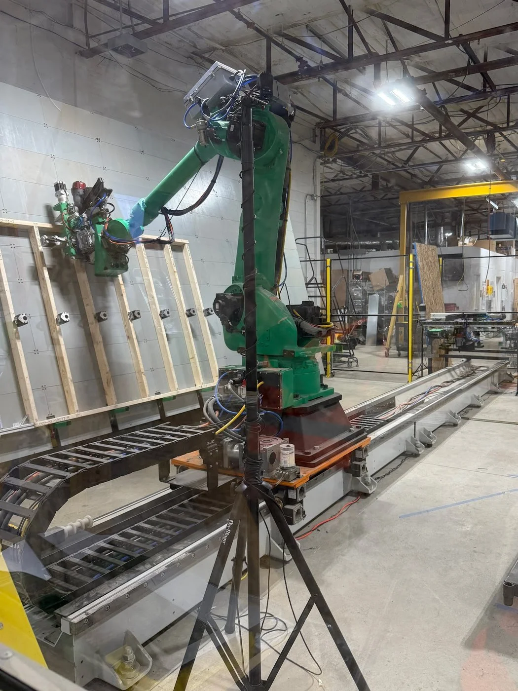 A green industrial robot arm on a floor rail works on a wood-framed wall panel inside a factory.