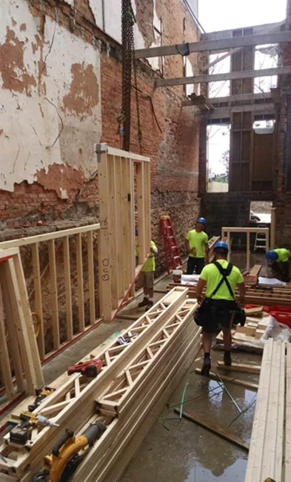 Construction workers in hard hats and high-visibility shirts frame new wood walls and floor trusses inside the stripped brick shell of an old rowhouse.