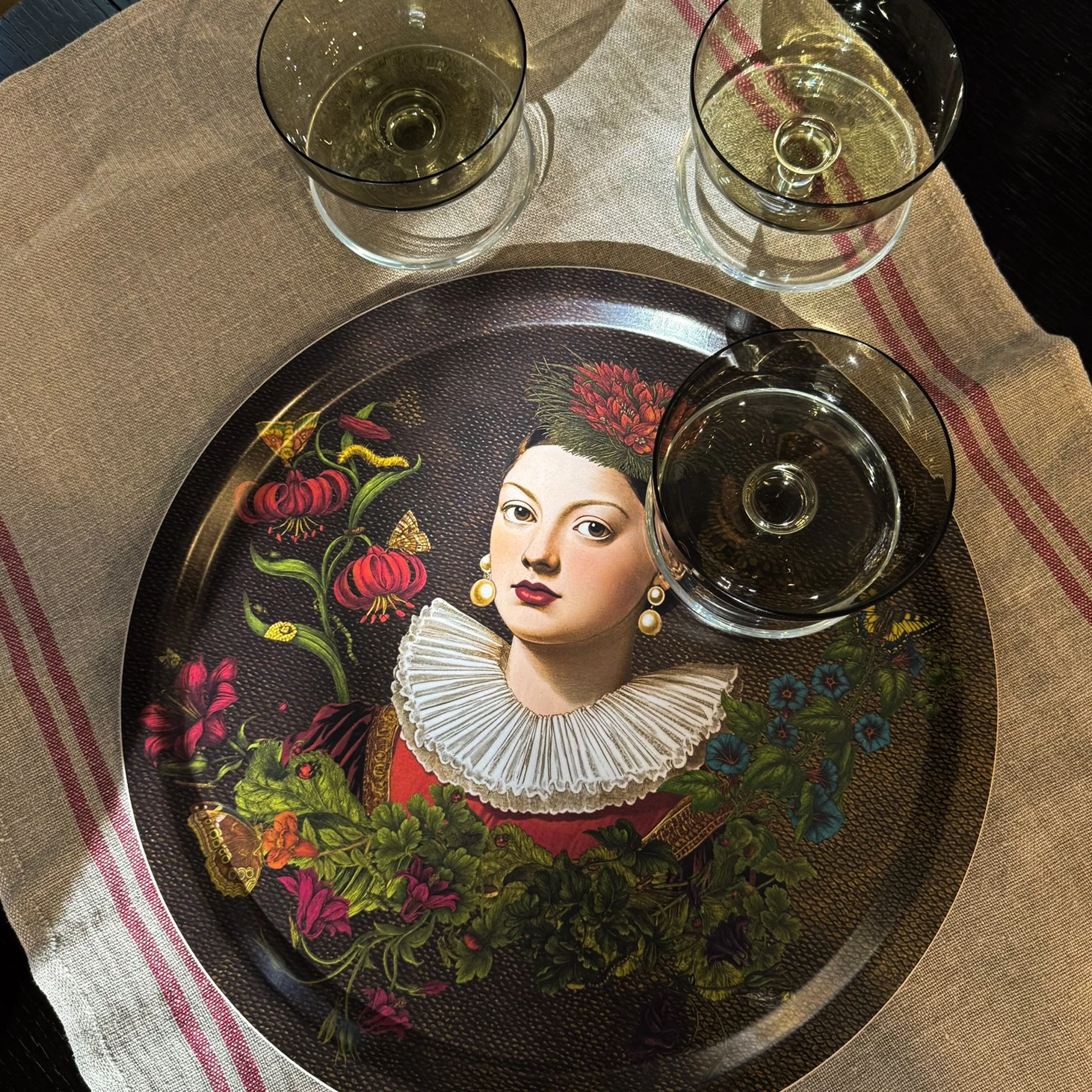 Round birchwood serving tray from the 'Flower Notes' collection. Features a surreal botanical illustration of a woman in a Renaissance ruff collar surrounded by lush flowers and insects. Designed by Laura Molina.