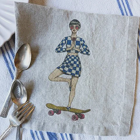 A napkin with an illustration of a woman balancing on a skateboard, wearing sunglasses, a checkered dress, and holding a book, with a spoon, knife, and fork placed beside it.
